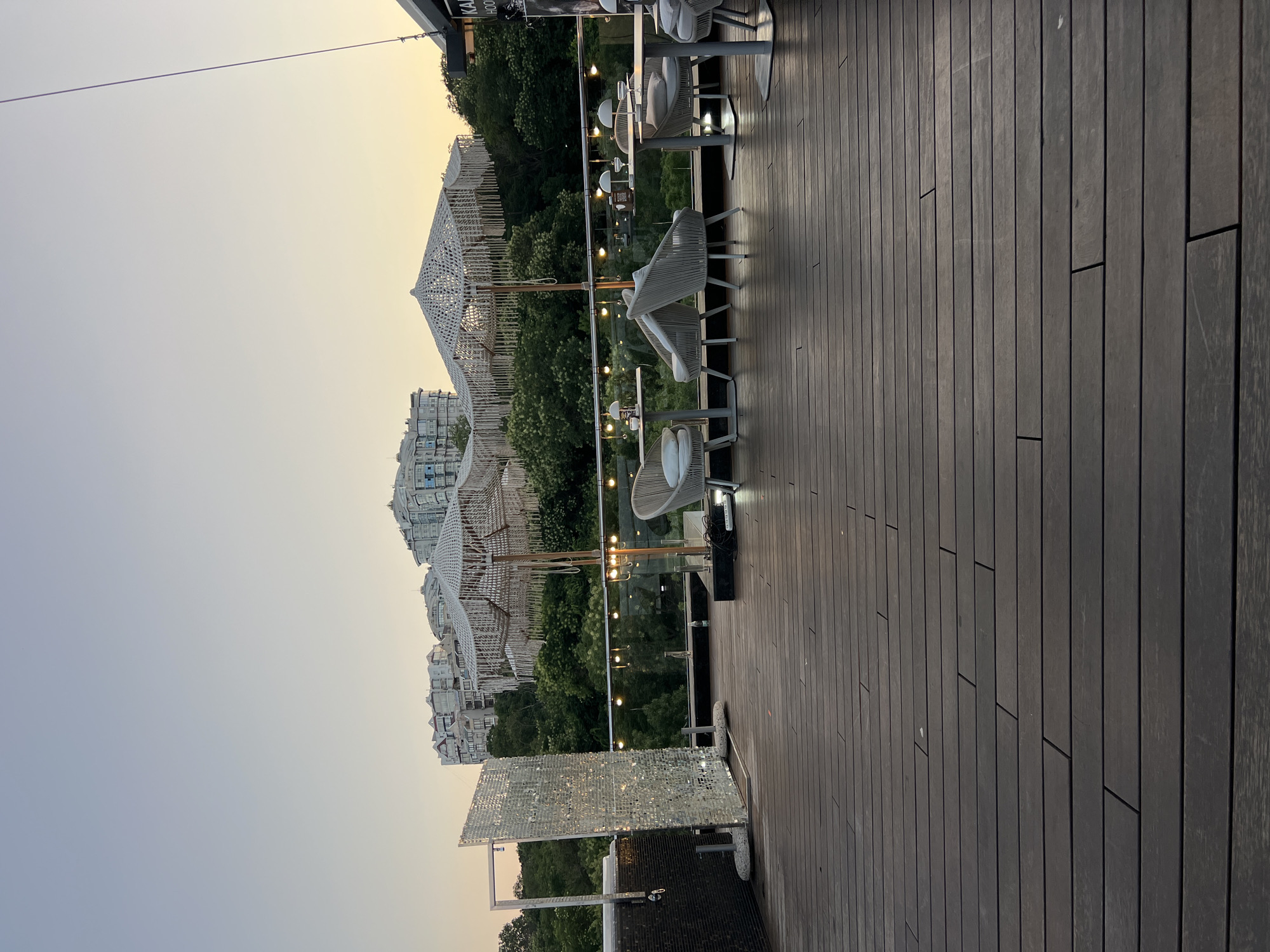 The image depicts a rooftop terrace in Odesa, Ukraine, at dusk. The terrace is furnished with several tables and chairs, some of which are occupied by unseen patrons. The flooring is made of dark wooden planks. In the background, a line of trees separates the terrace from a cityscape featuring modern residential buildings. Decorative macrame umbrellas are set up, and string lights are visible, suggesting an evening dining or relaxation setting. A large, reflective panel stands on the left side of the terrace, possibly for ambiance or as a decorative element. The sky is a gradient of pale blue and yellow, indicative of sunset.