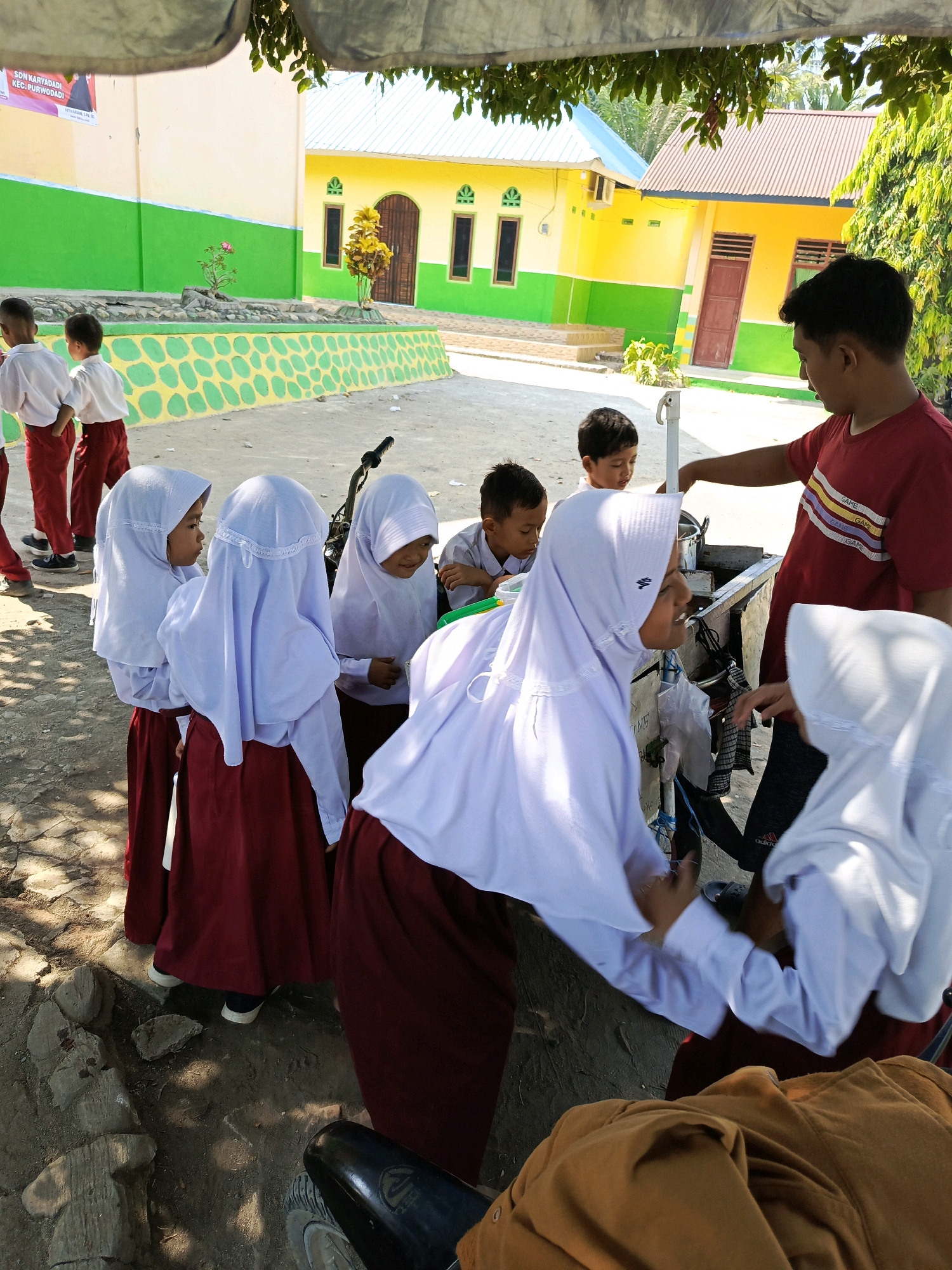 In the city of Karya Dadi, Indonesia, on a bright sunny day, a group of children, likely students, are gathered around a food cart. Several young girls in white hijabs and maroon skirts stand in a cluster, some facing each other in conversation. Two boys in white shirts and red pants are seen in the background, with one boy looking towards the food cart. A young man wearing a red t-shirt with colorful stripes is attending to the cart, interacting with the children. The environment appears to be an outdoor school area, with yellow and green buildings visible in the background, a decorative stone wall, and trees providing shade. The ground is a mix of dirt and gravel. A motorcycle is partially visible in the foreground, with its rear wheel and fender in view. The scene suggests a moment of social interaction and possibly a break or lunch period at a school.