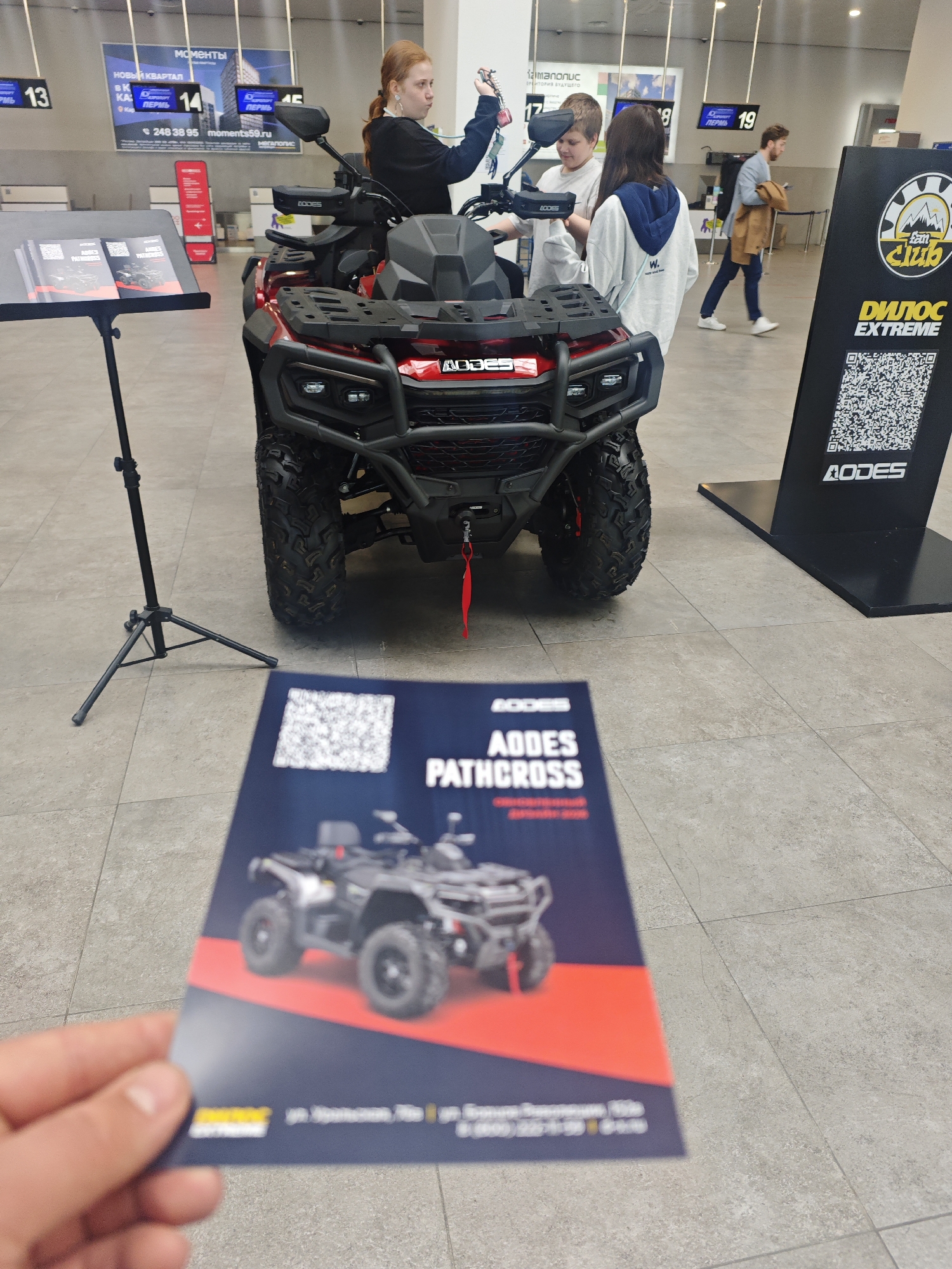 The image shows an AODES Pathcross all-terrain vehicle (ATV) displayed indoors, likely at an event or showroom. Three people, including a red-haired girl, are near the ATV. In the background, a man walks by. A stand with brochures and a sign with a QR code for "Дилос EXTREME" (Dilos Extreme) are also visible. A hand holds a brochure with the AODES Pathcross. Advertising for "новый квартал" (new quarter) "Пермь" (Perm) can be seen in the upper background. The location is indicated on the advertising banners as being in Perm, specifically "Мегаполис," which suggests the Bolshoye Savino International Airport.
