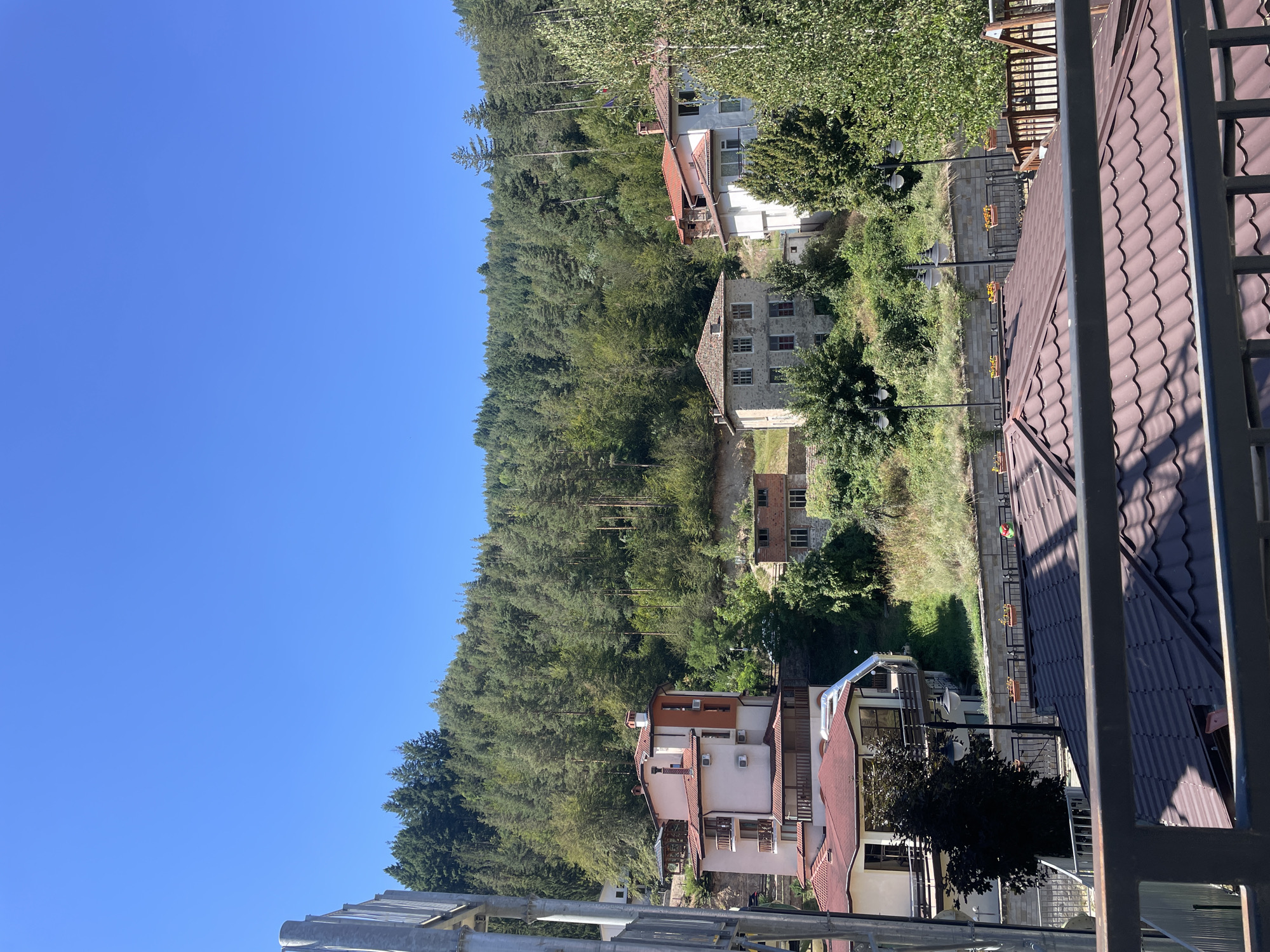 The image captures a view of buildings nestled on a hillside in the town of Banite, Bulgaria, under a clear blue sky. The foreground is dominated by the dark, patterned roof of a building and a metal railing, suggesting the vantage point is from a balcony or elevated structure.
Further up the slope, several houses are visible. One prominent building is painted in light pink and terracotta, featuring multiple balconies and air conditioning units. Adjacent to it, a smaller structure made of brick and stone with visible windows is partially overgrown with vegetation. Another house, constructed from stone blocks, displays multiple windows. In the background, a dense forest of pine trees covers the steep hillside.
The environment appears to be a residential area, characterized by the architecture of the houses and the natural landscape. The presence of trees, greenery, and the surrounding forest indicates a rural or semi-rural setting within the town of Banite. There are no people visible in the image. The lighting suggests it is daytime, with bright sunlight.
