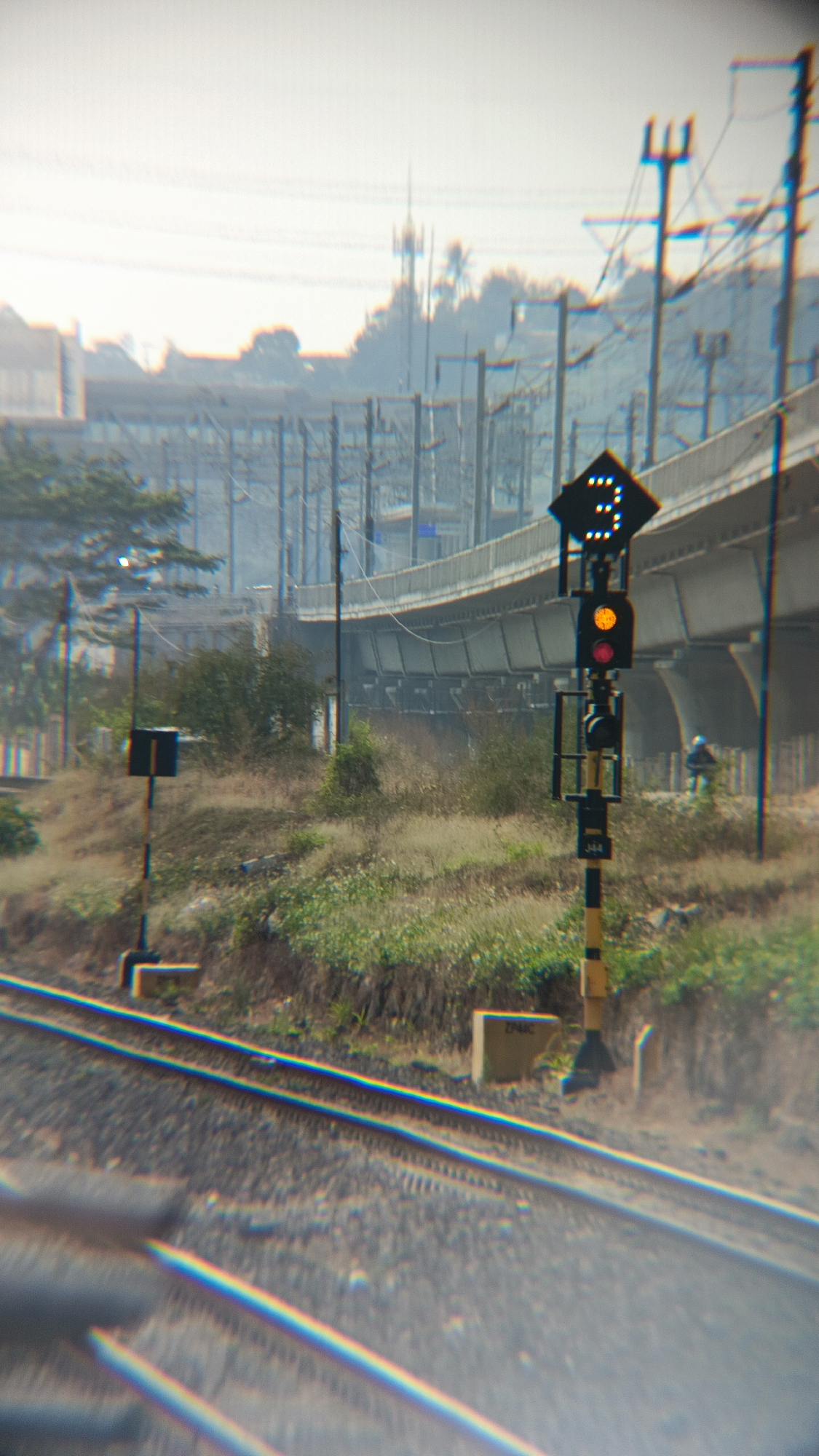 In Margajaya, Indonesia, a railway track stretches into the distance, flanked by dry, grassy embankments. A yellow and black striped pole stands to the left, topped with a dark square signal. To the right, a more elaborate railway signal post features a diamond-shaped sign with white illuminated dots arranged in a "3" formation. Below this, a traffic light displays a lit orange signal above a lit red signal. Further back, a concrete overpass with supporting pillars crosses the scene, with buildings and trees visible beyond. A solitary figure can be seen on the right, near the overpass. The sky is overcast.