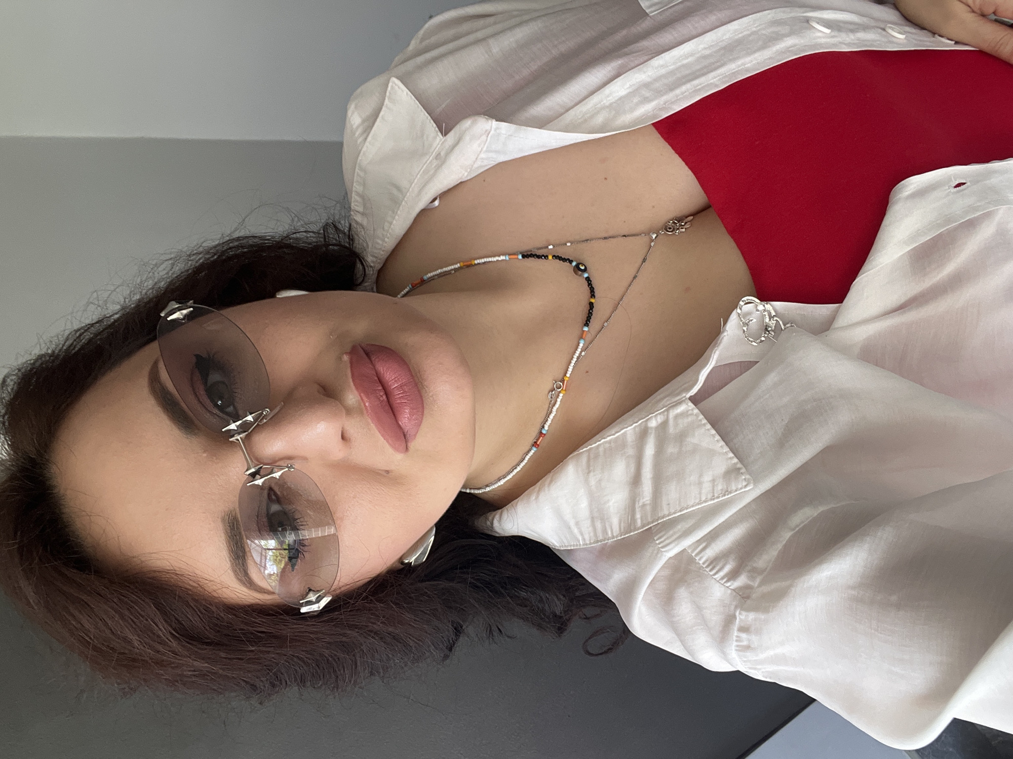A woman with dark brown hair and fair skin is pictured from the chest up. She is wearing a red top, a partially unbuttoned white shirt, silver earrings, and a beaded necklace with a dangling charm. She is also wearing sunglasses with a light brown tint and a silver frame. Her makeup includes dark eyeliner and lipstick. Her left ear is visible, and she appears to be wearing a white wireless earbud. The background is split between a dark gray wall on the left and a light gray wall on the right. The lighting suggests an indoor setting, possibly near a window.