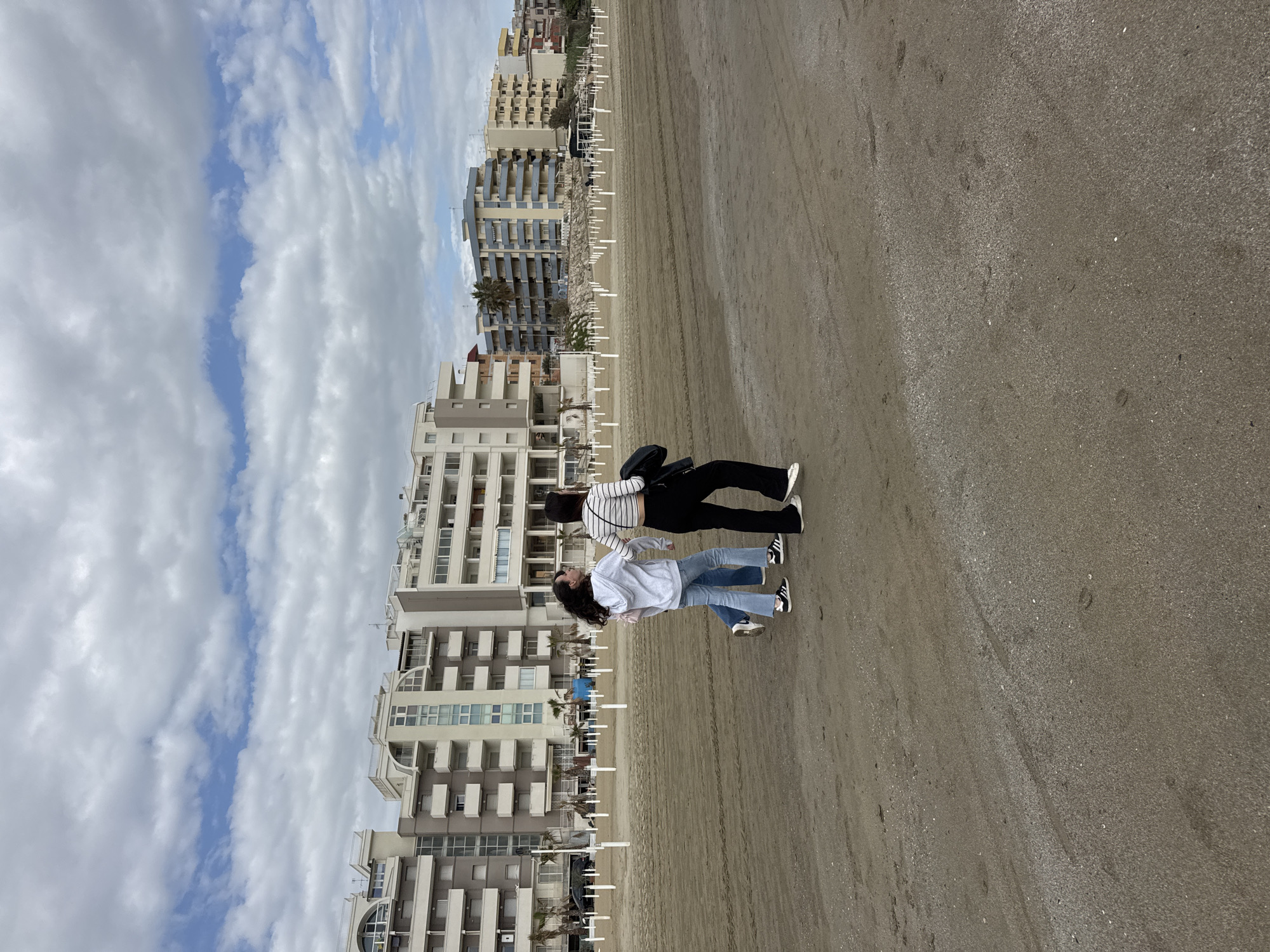 The image displays a coastal scene in Nettuno, Italy, featuring two young women on a wide sandy beach under a partly cloudy sky. The original image is rotated 90 degrees clockwise; when viewed upright, the sky occupies the upper portion, the beach the lower and right-hand side, and buildings line the left.
The two women are positioned towards the center-left of the frame on the sand. The woman on the left wears a light-colored sweatshirt, blue jeans, and sneakers, looking slightly upwards. The woman on the right, standing slightly behind her, is dressed in a striped long-sleeved top, black trousers, and sneakers, with a black shoulder bag, and appears to be looking towards the first woman.
Behind the women, a row of multi-story buildings, consistent with hotels or apartments, stretches along the beachfront. These structures exhibit various architectural styles, with many balconies and windows. Interspersed between the buildings and the sand are numerous white, thin, pole-like structures, typical of beach establishments for sun loungers or umbrellas. A few palm trees can be seen among the buildings.
The beach is extensive and appears to be packed sand, with visible tracks and subtle texture variations. The sky is mostly cloudy with scattered patches of blue, indicating a daytime setting that is not entirely sunny. The overall impression is one of a relaxed day at a coastal resort. No visible text or discernible brands are present in the image.