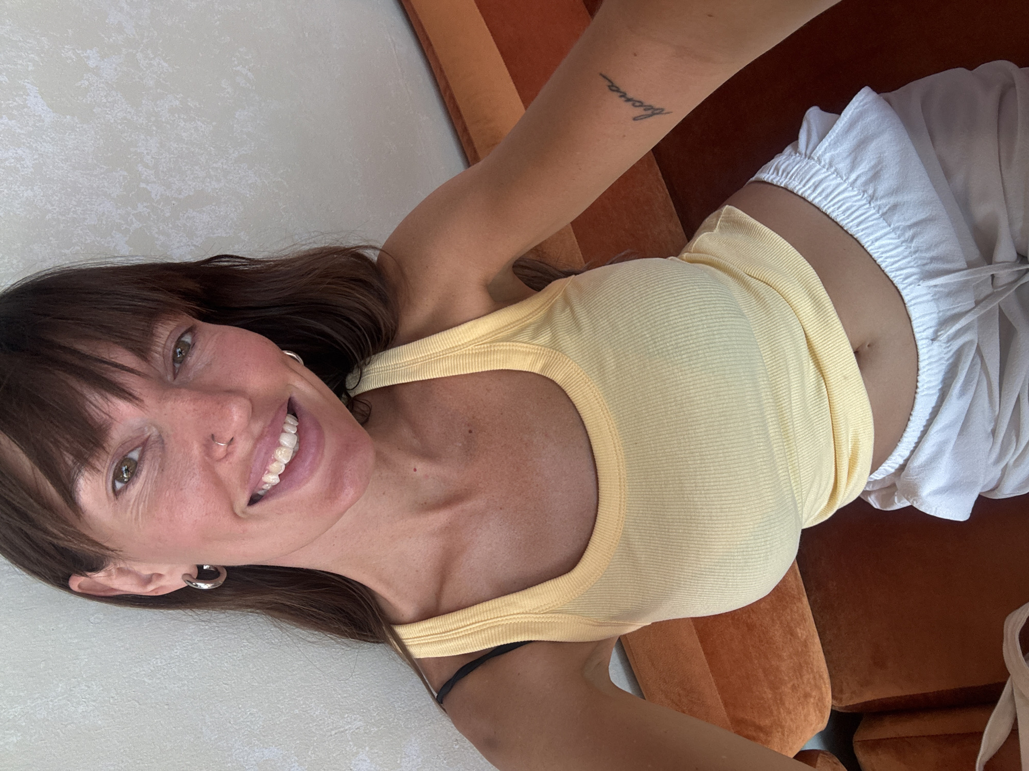 A woman is seen in an upward-angled selfie, lying on what appears to be an orange couch. She has brown hair with bangs, light skin, and is smiling. She is wearing a yellow tank top and white drawstring shorts. A tattoo is visible on her left arm, and she has a nose ring and hoop earrings. The location is not discernible from the image.