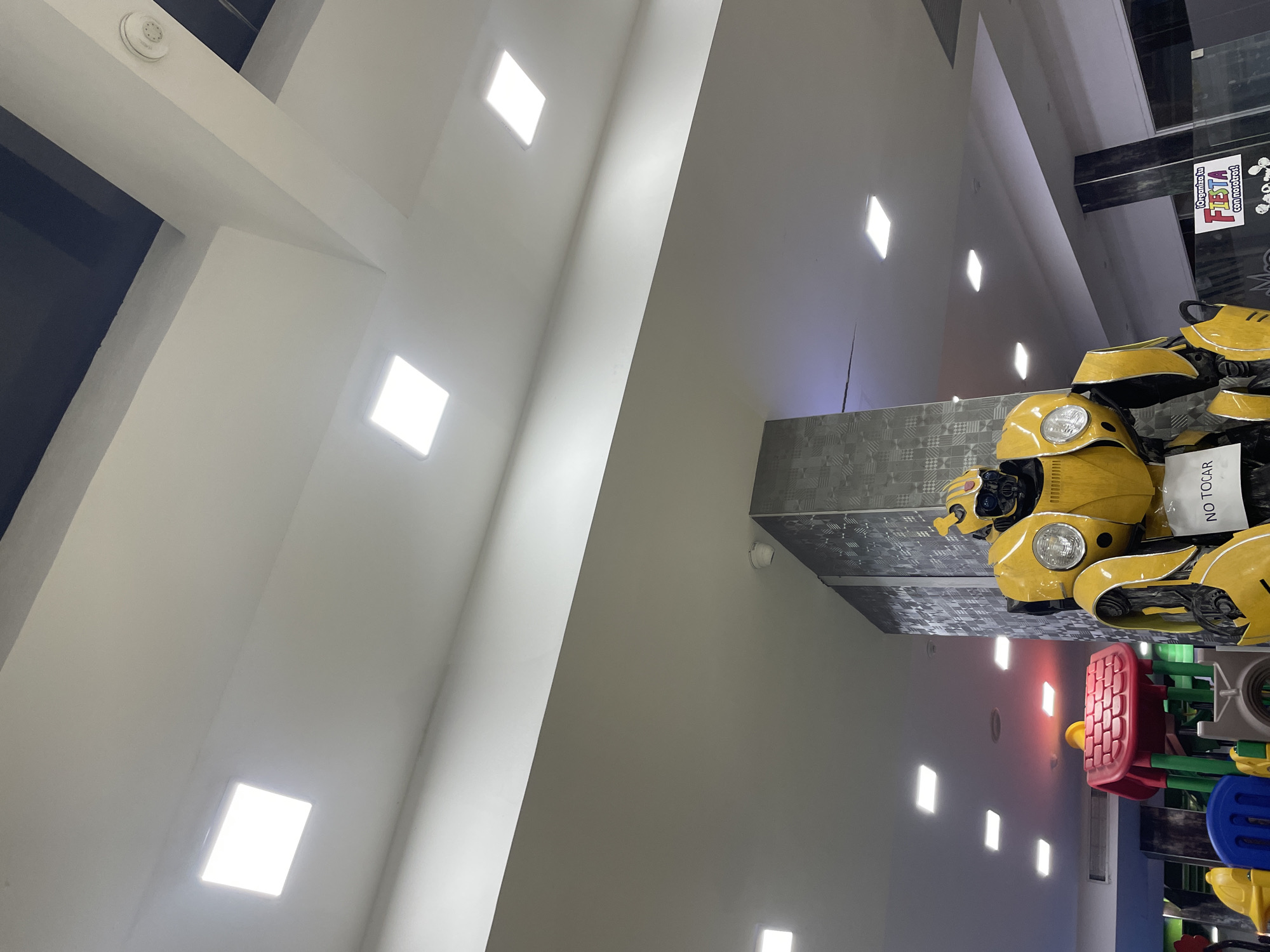 This image depicts an indoor scene with a large, yellow, robot figure resembling Bumblebee from the Transformers franchise. The robot is positioned in the foreground and is adorned with a sign that reads "NO TOCAR," meaning "DO NOT TOUCH" in Spanish. To the left of the robot, a colorful children's play structure is visible, featuring red, blue, and green components. The environment appears to be a commercial or entertainment space, with a white ceiling populated by numerous square, illuminated light panels. A decorative pillar with a gray, patterned texture stands behind the robot. The overall impression is that of an indoor amusement area or a store display. The specific location in El Valle del Espíritu Santo, Venezuela, is not visually indicated in the image itself.
