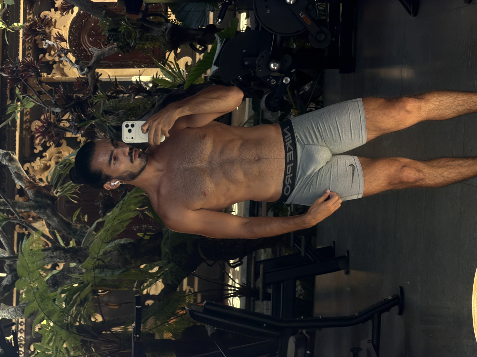 A shirtless man in gray athletic shorts with "NIKE PRO" branding stands inside, holding a smartphone and appearing to take a selfie. He has a beard, dark hair pulled back, and an earbud in his left ear. The background features tropical plants and what appears to be elaborate wooden carvings, suggesting an ornate interior. Behind the man, a piece of exercise equipment is visible. This scene is possibly set in a fitness area or gym located in North Kuta, Indonesia.