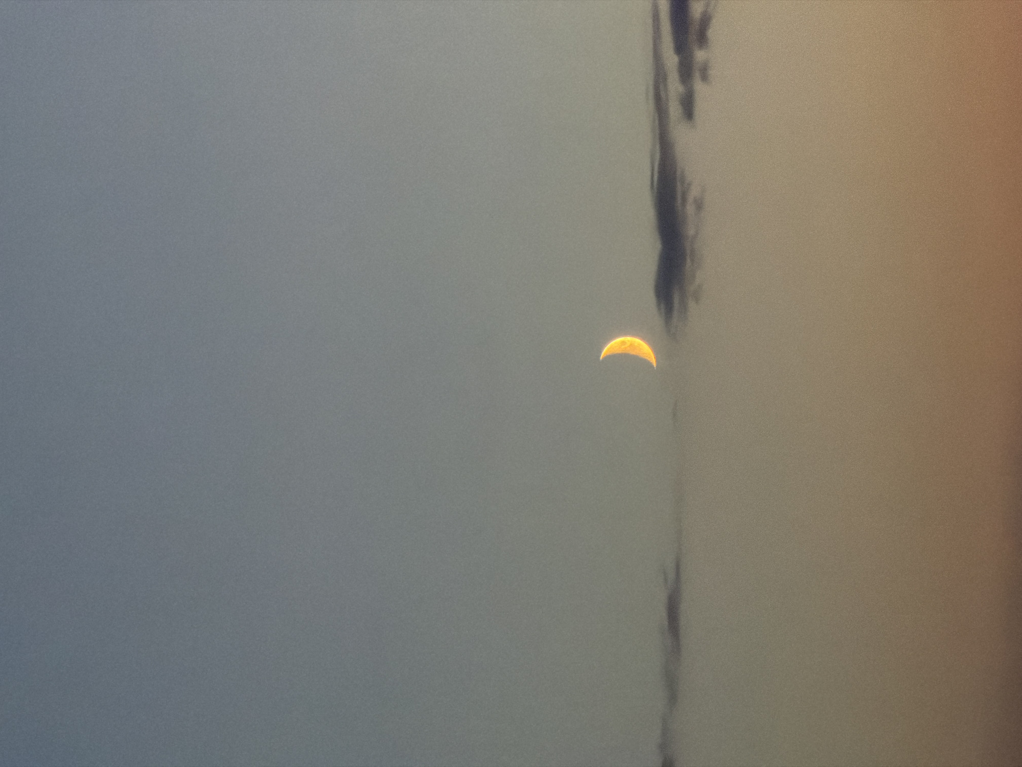 The image captures a crescent moon in a twilight sky. The moon is illuminated with a warm, golden glow and is positioned to the left of center. Below the moon, a thin band of dark clouds stretches horizontally across the frame. The sky transitions from a soft blue at the top to a gradient of orange and yellow towards the bottom, suggesting the lingering light of sunset. The image does not contain any elements that would indicate a specific location in Saint Petersburg, Russia, or any people, actions, or interactions. The scene is purely naturalistic, focusing on the celestial body and atmospheric conditions.