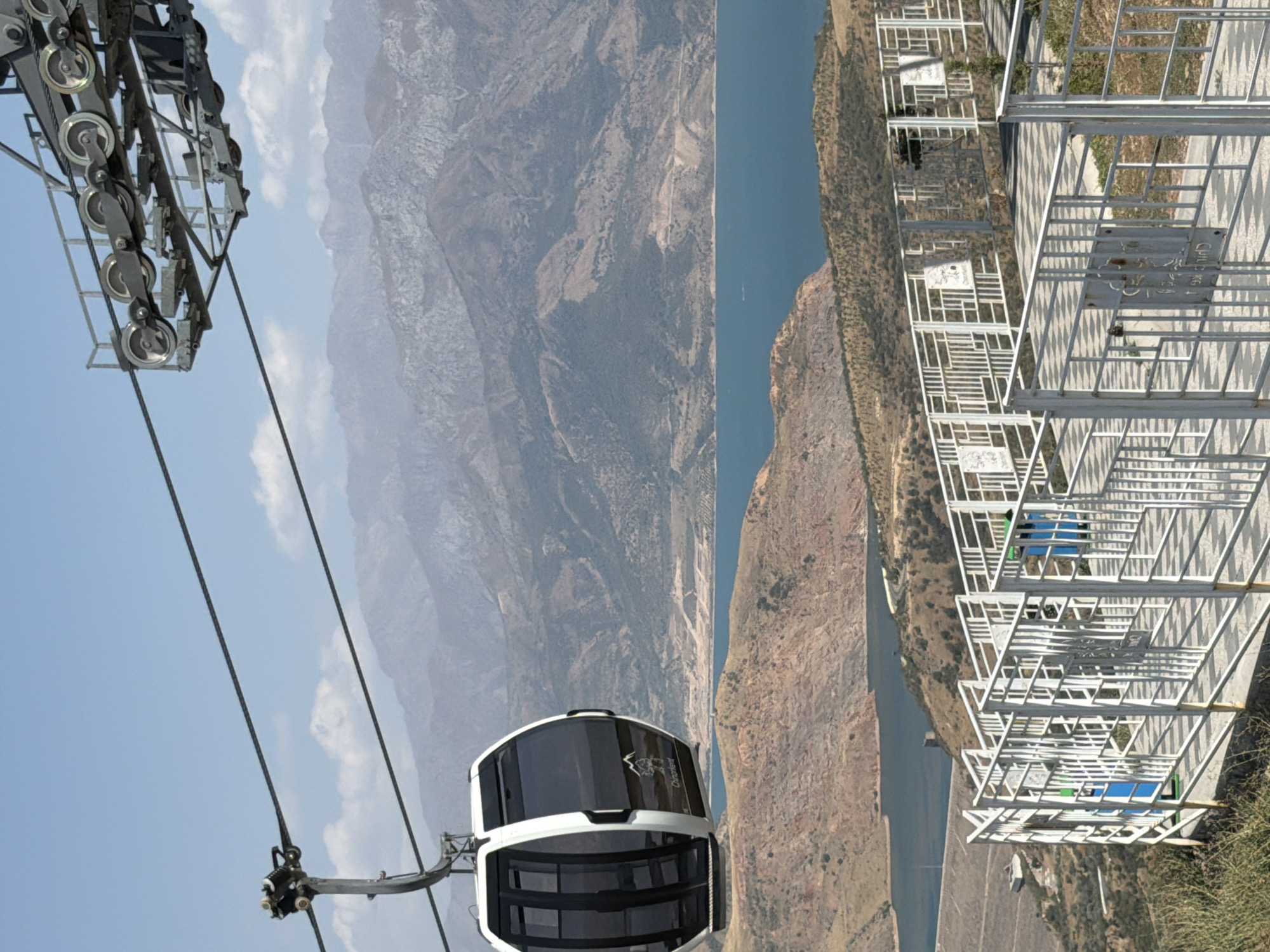The image captures a view from a cable car station overlooking a reservoir and mountainous terrain. In the foreground, a white metal railing encloses a paved walkway, with two blue trash receptacles visible. Several signs are mounted on the railing, some displaying images and text. A modern cable car cabin, white with black windows and a logo, is suspended from a cable overhead. In the background, a large body of blue water, likely a reservoir, is visible, with arid, mountainous landscapes stretching towards the horizon. The overall environment is dry and sunny, with a clear blue sky. The scene is located in the vicinity of Charvak, Uzbekistan, a popular tourist destination known for its reservoir and surrounding natural beauty.