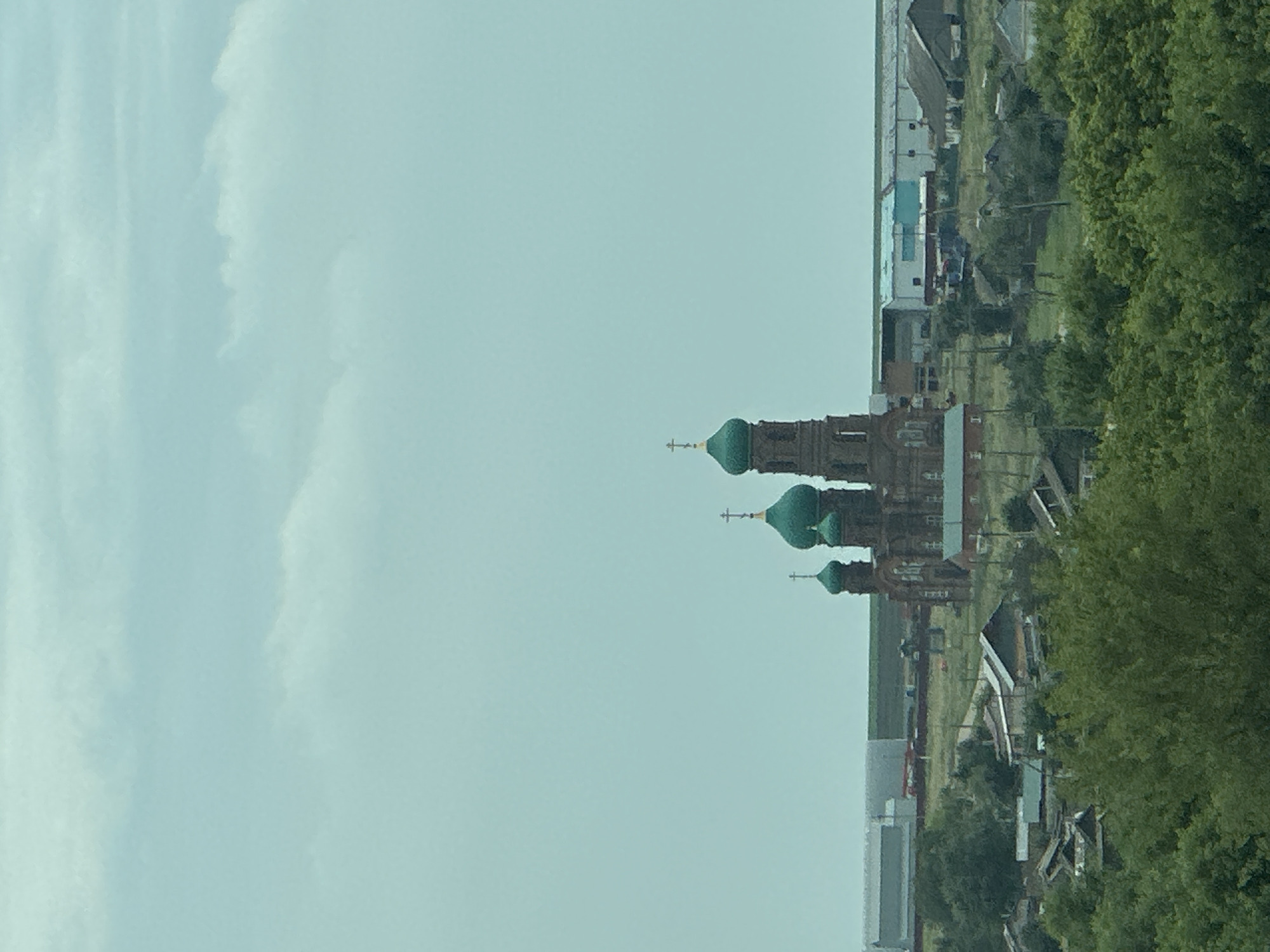 An elevated, medium-shot captures a view of a village in Yekaterinovka, Russia, with a prominent brick church with green domes in the background. The sky is overcast and a pale blue, dominating the upper half of the frame. Below the sky, a hazy line separates the sky from the landscape. In the mid-ground, the church stands out with its brick facade and three distinct green-domed towers, each topped with a cross. To the left and right of the church, several low-rise buildings with grey and blue roofs are visible, suggesting an industrial or agricultural area. The foreground is filled with lush green trees, obscuring the lower portion of the village buildings. The overall impression is one of a rural settlement under a cloudy sky, with the church as a central architectural element. There are no people or animals visible in the scene.