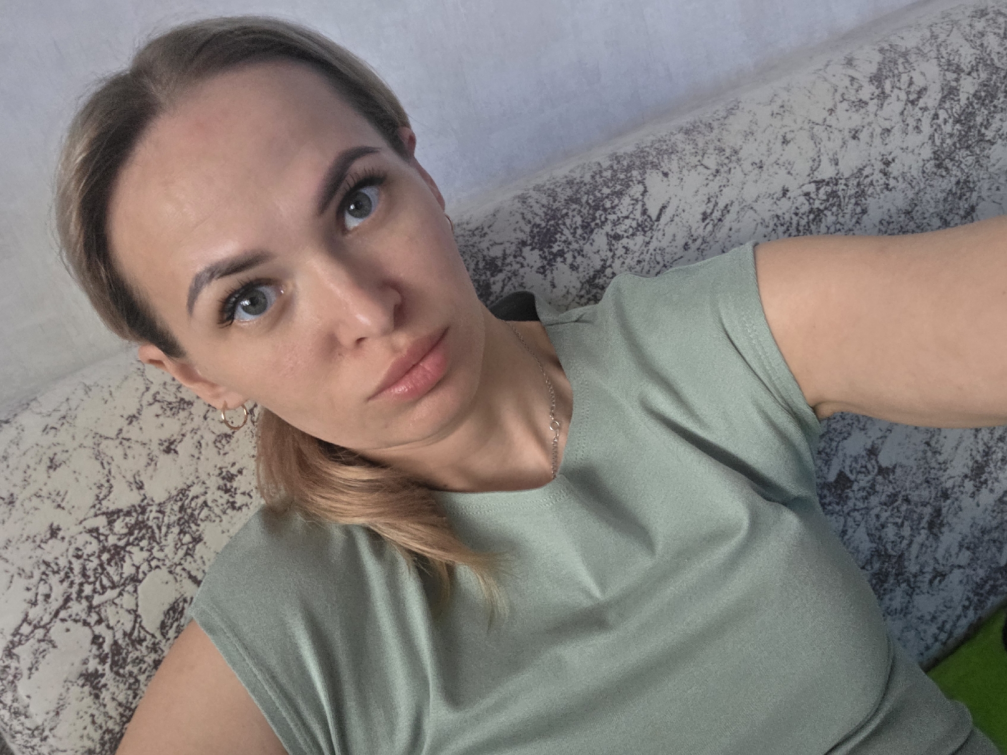 A selfie of a fair-skinned woman with light brown hair pulled back in a low ponytail, wearing a light green t-shirt and a delicate silver necklace. She has green eyes and is looking directly at the camera with a slightly serious expression. She is wearing small gold hoop earrings. The background suggests she is sitting on a patterned sofa or couch. The image was likely taken indoors in the city of Chaikovsky, Russia.