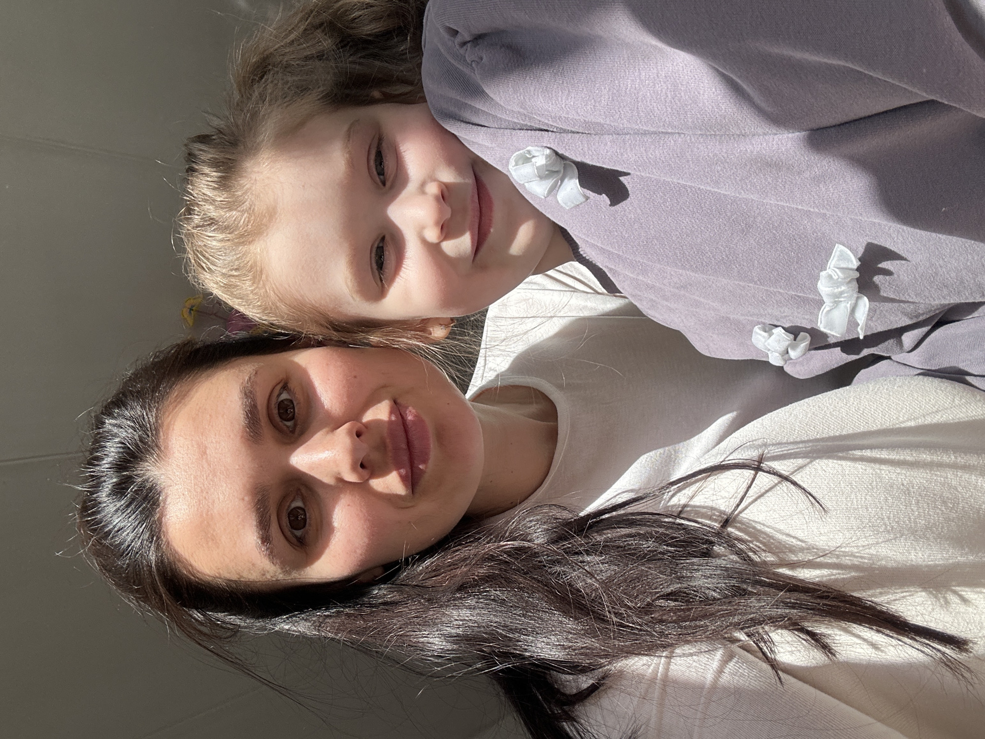 A selfie photograph features a woman and a child. The woman has dark hair and brown eyes, and she is wearing a light-colored shirt. The child, presumed to be her daughter, has blonde hair and is wearing a lavender-colored garment with small white bows. The background suggests an indoor setting with natural light. It's impossible to verify the location as Chaikovsky, Russia, without additional information.