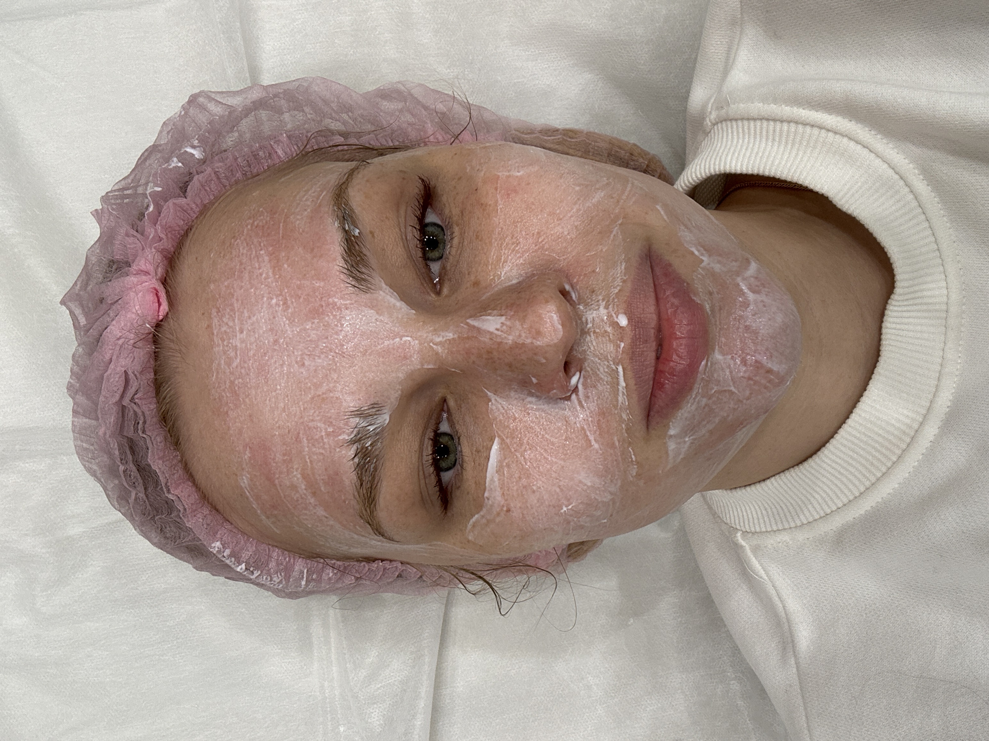 A light-skinned person wearing a pink disposable hair covering lies on a white sheet. The person's face is covered with a white, possibly creamy, substance. The person appears to be wearing a white sweatshirt. There are no discernible location cues to identify Rusanovo, Russia.