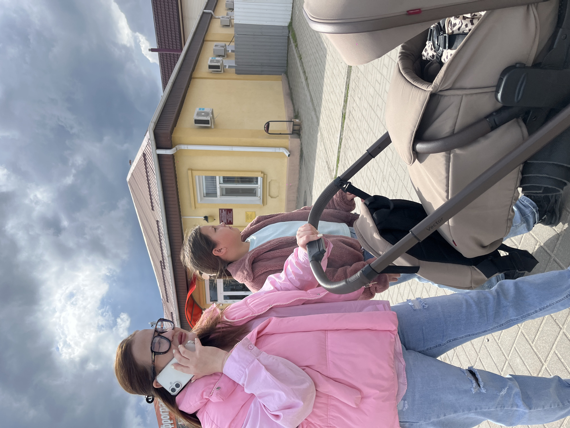 A photo shows two people on a paved street in Novopokrovskaya, Russia, next to a light yellow building with a brown roof. The person closest to the camera wears glasses, a pink jacket, and ripped jeans while talking on a white phone. The second person wears a brown jacket and blue jeans, holding onto the handle of a beige stroller. The sky is cloudy.