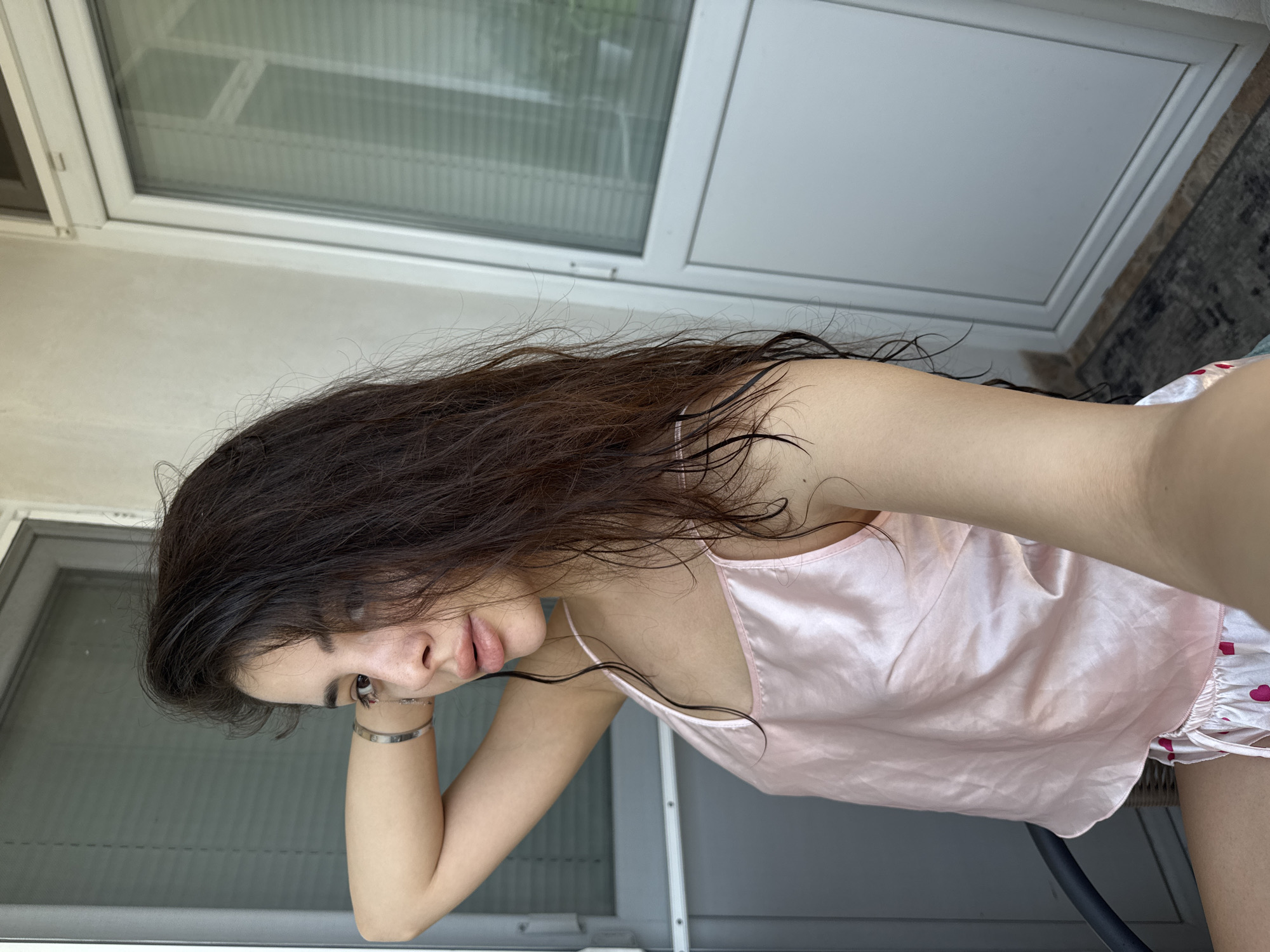 A female subject is depicted in a self-portrait taken in Plovdiv, Bulgaria. She is positioned with her body slightly angled to the viewer's right and her head turned towards the camera, maintaining direct eye contact. Her long, dark brown hair, which appears wet, is styled over her shoulders, with some strands draped across the left side of her face. She wears a light pink, shiny camisole top with thin straps, revealing her upper torso. Her left arm is bent and raised, with her hand positioned behind her head, showcasing a silver-colored bracelet on her wrist. Her lips are full and slightly parted, revealing no teeth. The background consists of architectural elements, suggesting an interior or semi-enclosed outdoor space. To the left is a dark gray framed door with horizontal slats visible through a panel. To the right, a white framed door with a large glass panel and horizontal blinds is visible. The wall above these structures is light-colored and unadorned. A section of a dark-colored textured floor covering or mat is visible at the bottom right, adjacent to the white door's threshold.