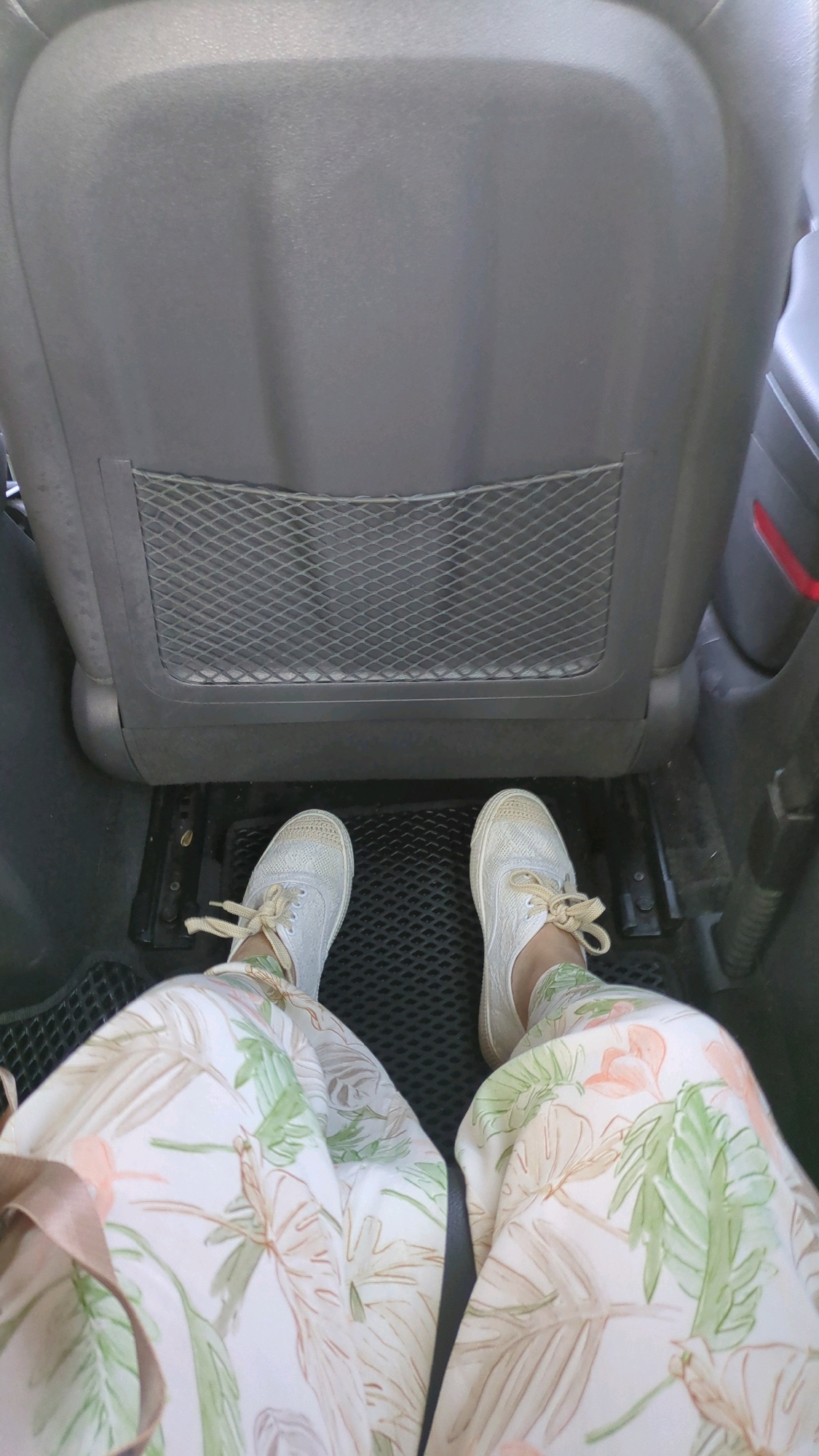 The image shows a person's legs and feet in a car. The person is wearing white sneakers and pants with a floral pattern of green and peach leaves and flowers. They are sitting in the back seat of the car, and the back of the car seat in front of them is visible, with a mesh pocket. The floor of the car has a black rubber mat. The image does not provide any information about the location in Chișinău, Moldova.