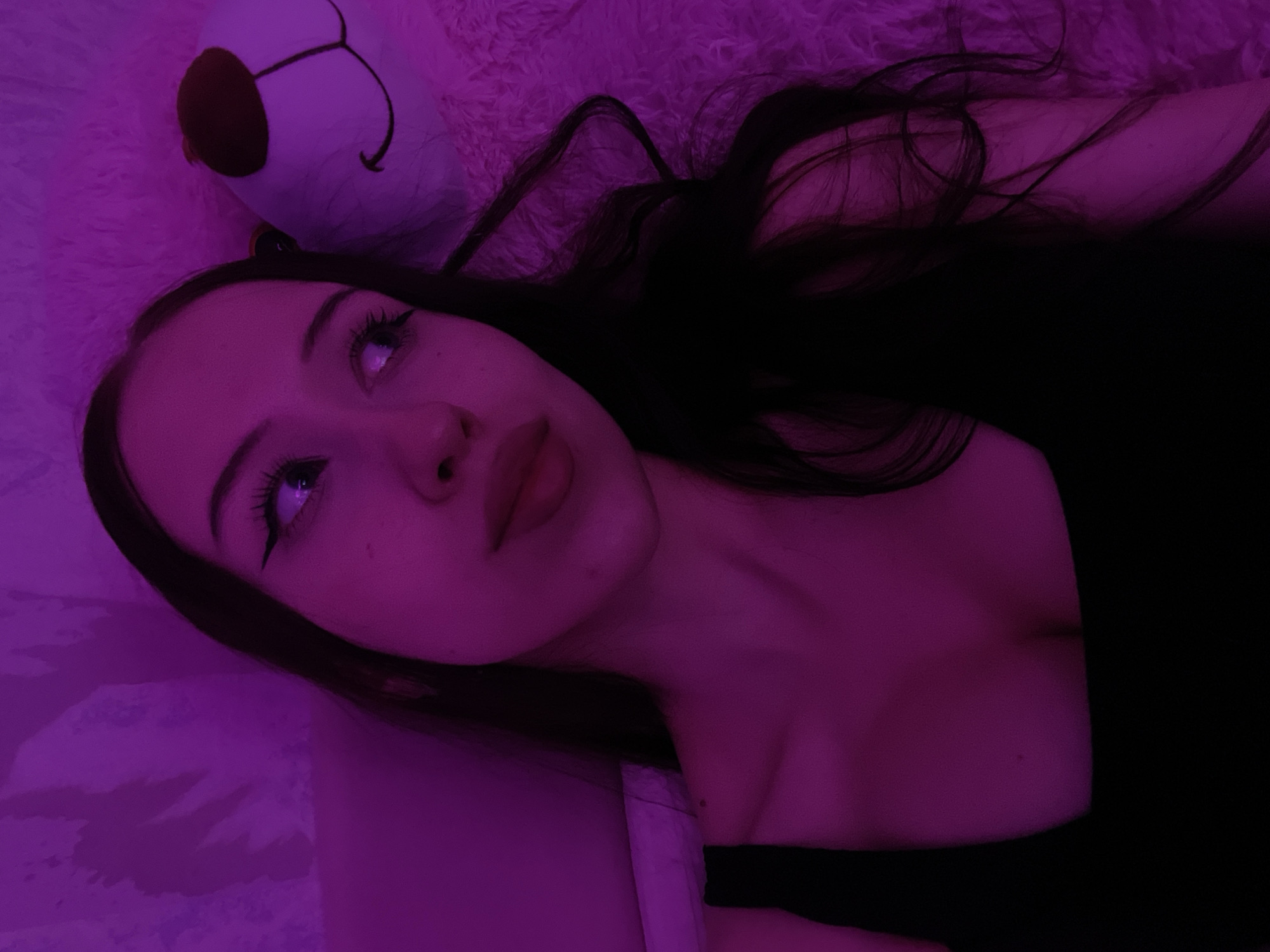 A young woman with long, dark hair and noticeable eyeliner is illuminated by purple lighting. She is lying down on a fluffy pink surface, possibly a blanket or a stuffed animal, as evidenced by the texture. To her right, the face of a large, white teddy bear with brown ears and a black stitched smile is visible. The woman is wearing a black tank top. The background appears to be a textured wall. No specific location in Votkinsk, Russia, can be determined from the image.