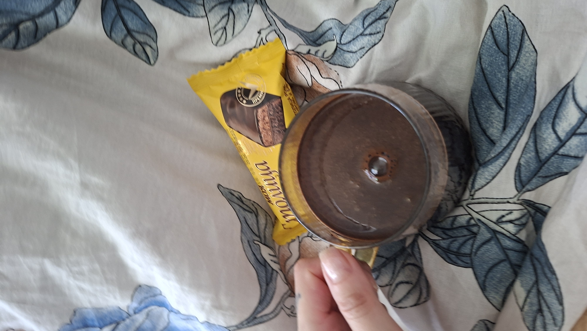 A hand with a manicured nail is seen reaching towards a glass of dark liquid on a patterned white surface. Adjacent to the glass is a yellow candy wrapper with Russian text and an image of a chocolate confection. The surface appears to be fabric with a repeating motif of blue leaves. The overall scene suggests a casual moment, possibly a snack break. The specific location in Myadzyel, Belarus cannot be determined from the image.
