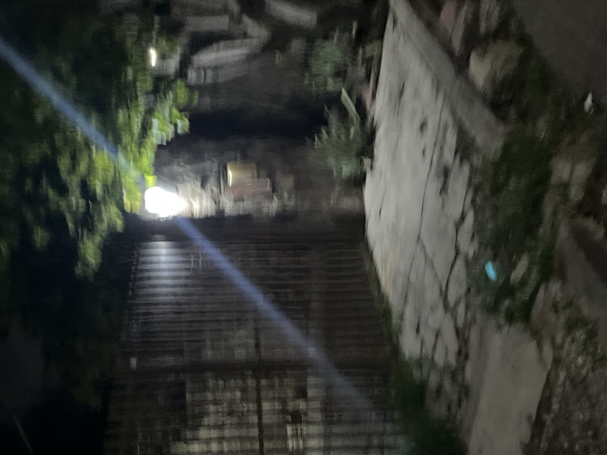 The image depicts an outdoor scene at night, likely in Santa Ana, Venezuela. A corrugated metal fence dominates the left side of the frame. To the right of the fence, a stone wall is visible, illuminated by a bright light source originating from above and to the left, casting a prominent diagonal beam of light across the image. In front of the stone wall, there is a patch of light-colored paving stones. Further to the right, an area of dense, dark vegetation is present, with some lighter green foliage interspersed. The ground in the foreground appears to be a mix of gravel and grass, with some debris scattered about. The overall lighting is dim, suggesting nighttime conditions, with the bright light source creating significant contrast and lens flare. The scene is devoid of any people or discernible activity.