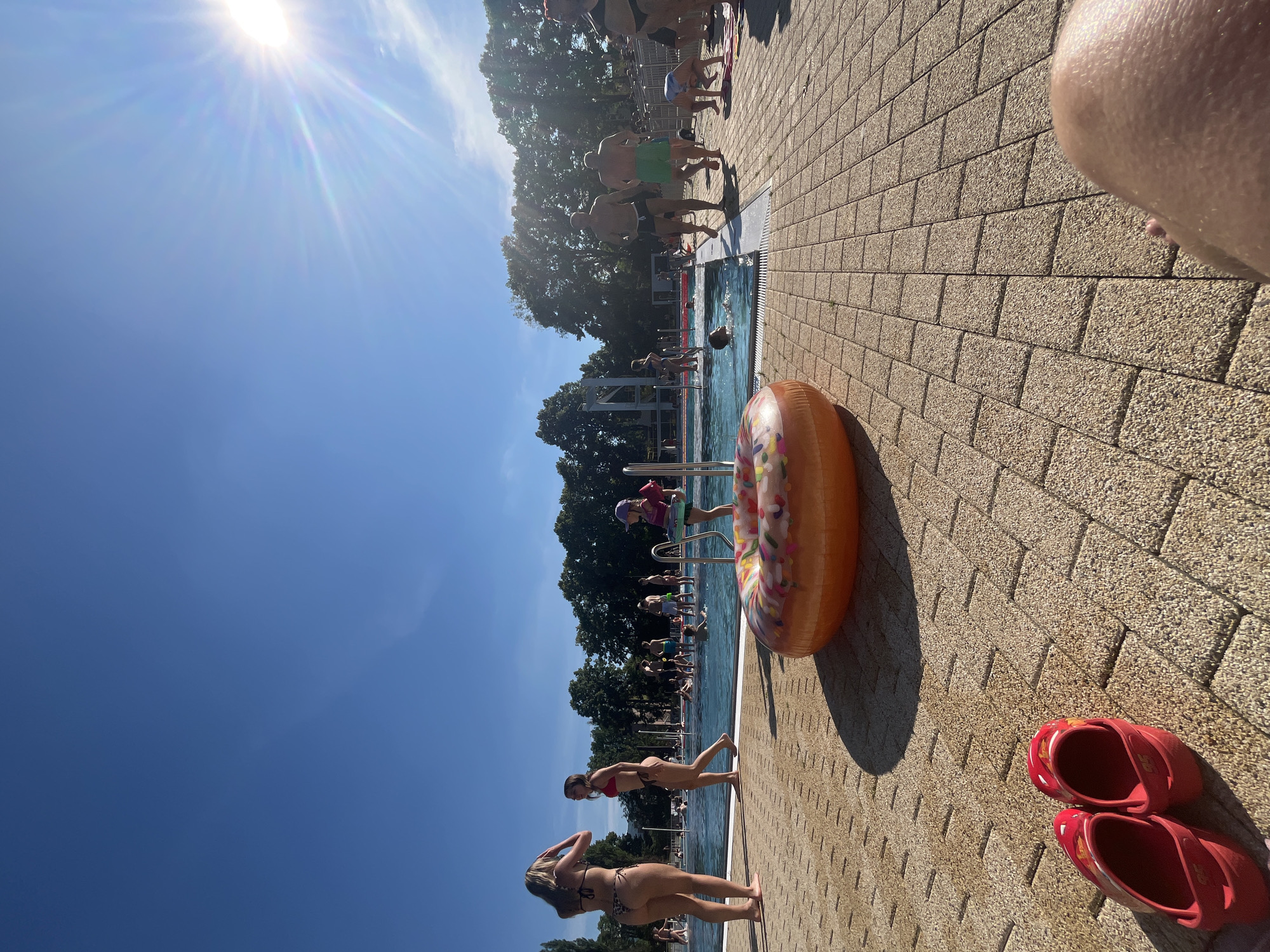 An outdoor swimming pool area is depicted on a sunny day. In the foreground, a large orange inflatable donut float rests on the tan brick pavement. To the left of the float, a pair of red clogs are placed next to each other. Several people are visible throughout the scene, many of whom are in or near the pool. Two women in bikinis stand by the edge of the pool on the left. A young girl in a purple hat and pink arm float is standing on the pool steps. In the middle ground, several individuals are swimming in the clear blue water, while others are standing or walking near the poolside. The background features a line of lush green trees and a bright blue sky with the sun prominently shining in the upper right corner, creating lens flare. The overall impression is a lively and recreational atmosphere at a public pool.
