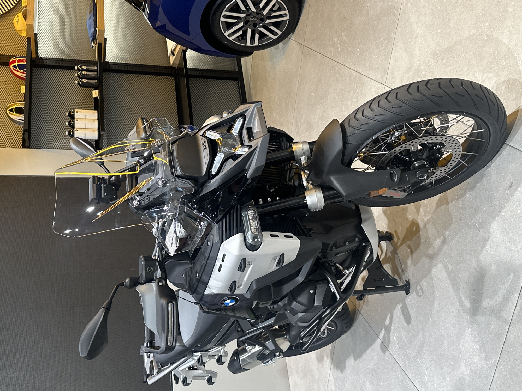 The image depicts a BMW R 1250 GS motorcycle displayed indoors, likely in a showroom or exhibition. The motorcycle is predominantly black and silver with a transparent windscreen. The front wheel is prominently featured, showing a knobby tire suitable for off-road or adventure riding.
Behind the motorcycle, there is shelving with various items, including what appears to be motorcycle helmets and other accessories. Above the motorcycle is the tire of a Mini Cooper.
Based on the presence of these items, the location is most likely a BMW or multi-brand automotive and motorcycle dealership or a promotional event space. Without more visual clues, pinpointing an exact location in Bangkok is not possible.