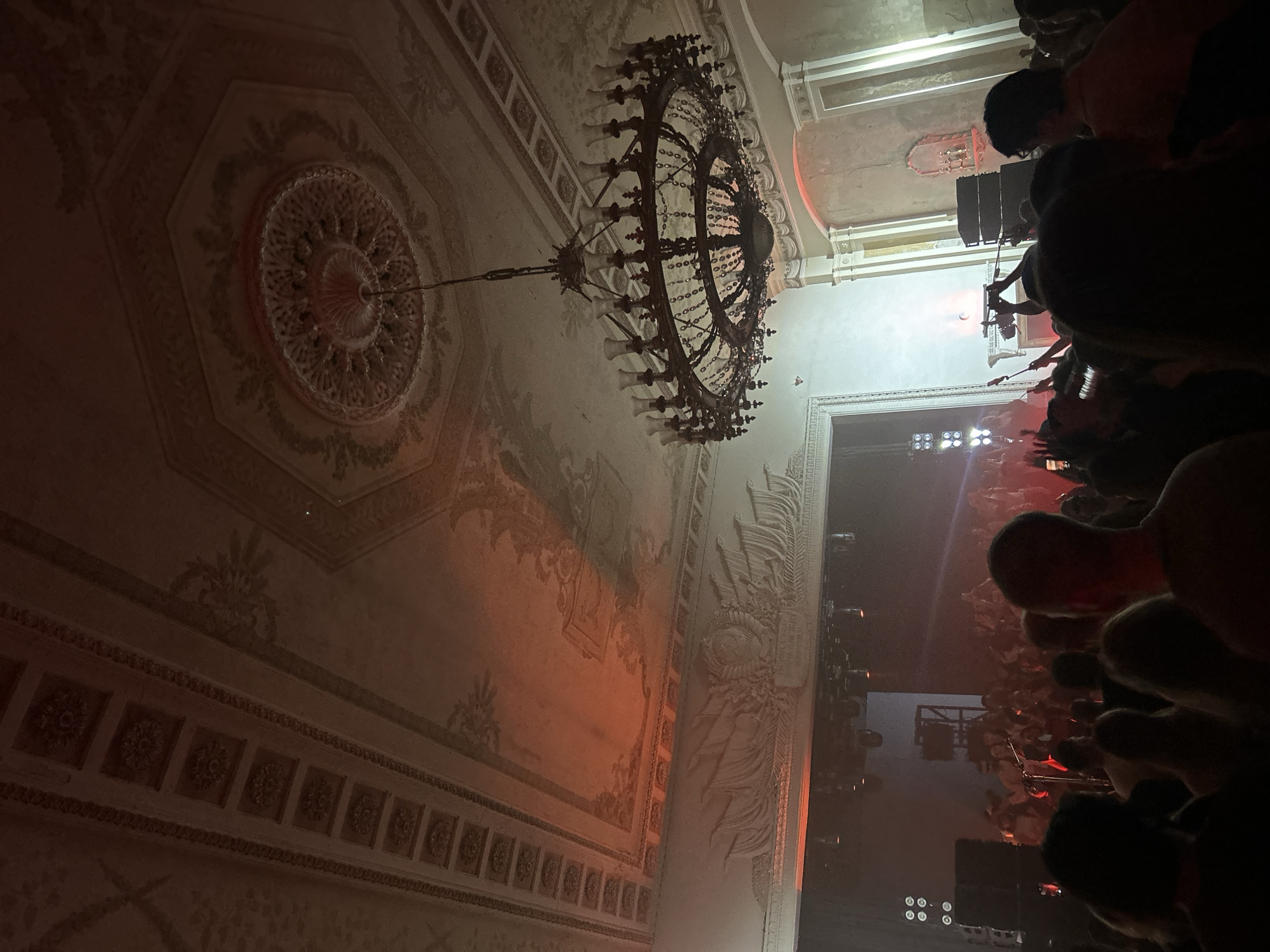 The image shows a crowded concert hall. The ceiling features ornate plasterwork and a large chandelier. In the foreground, audience members stand close together, obscuring the stage. The lighting suggests a performance is in progress. The location is likely in Moscow, Russia, given the architectural style.