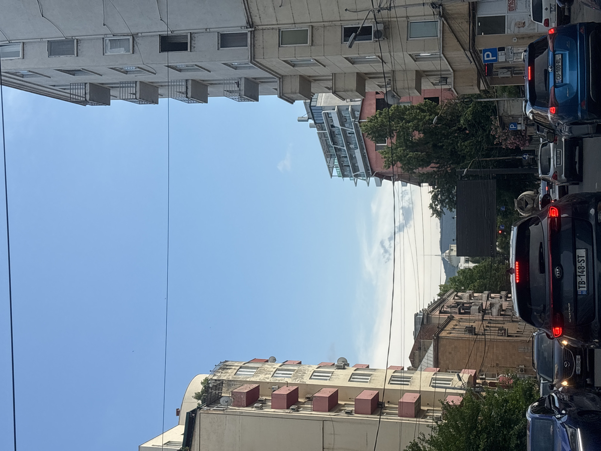 The media file shows a street scene in Tbilisi, Georgia, likely during the day. The perspective is from a vehicle, as indicated by the view of the road and other cars. The image is oriented vertically.
The scene is dominated by buildings on either side of the street. On the left, a tall, light-colored apartment building with many windows and protruding balconies is visible. On the right, a mix of buildings is present, including a pink building with trees in front, and further down the street, a taller, older-looking building with many windows and external air conditioning units. Traffic is visibly backed up, with several cars in a line. One car, a blue Kia Sportage, has a license plate reading "CU-100-RL" and "TB-148-ST". Another car has a license plate "HMU-325-60". Blue parking signs with a "P" are visible on poles.
The sky is clear and blue with some wispy clouds. The overall impression is of a typical urban street with moderate traffic. The time of day appears to be daytime, possibly late morning or afternoon, given the bright sunlight. The weather seems to be clear and pleasant. There are no people clearly visible in the image. The focus is on the architecture and the streetscape.