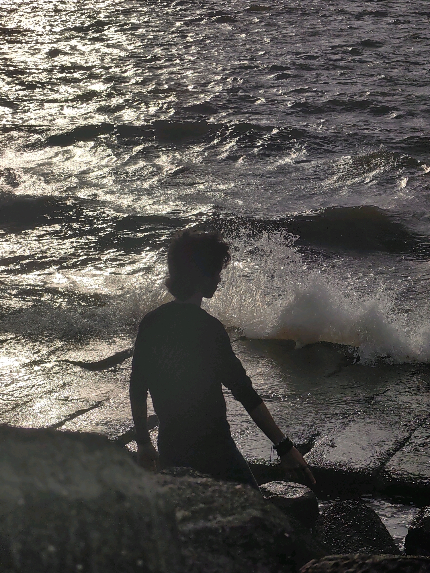 A silhouette of a person with curly hair sits on rocks by the ocean. The person is wearing a dark long-sleeved shirt and has a bracelet on their right wrist. Their right hand is extended towards the water, with fingertips touching a rock. Waves crash against the shore, creating white foam. The sun is setting, casting a shimmering reflection on the water. The image is taken from a low angle, looking up at the person and the ocean. This scene could be located at Marine Drive in Mumbai, India, a popular promenade known for its ocean views and rocky shorelines.