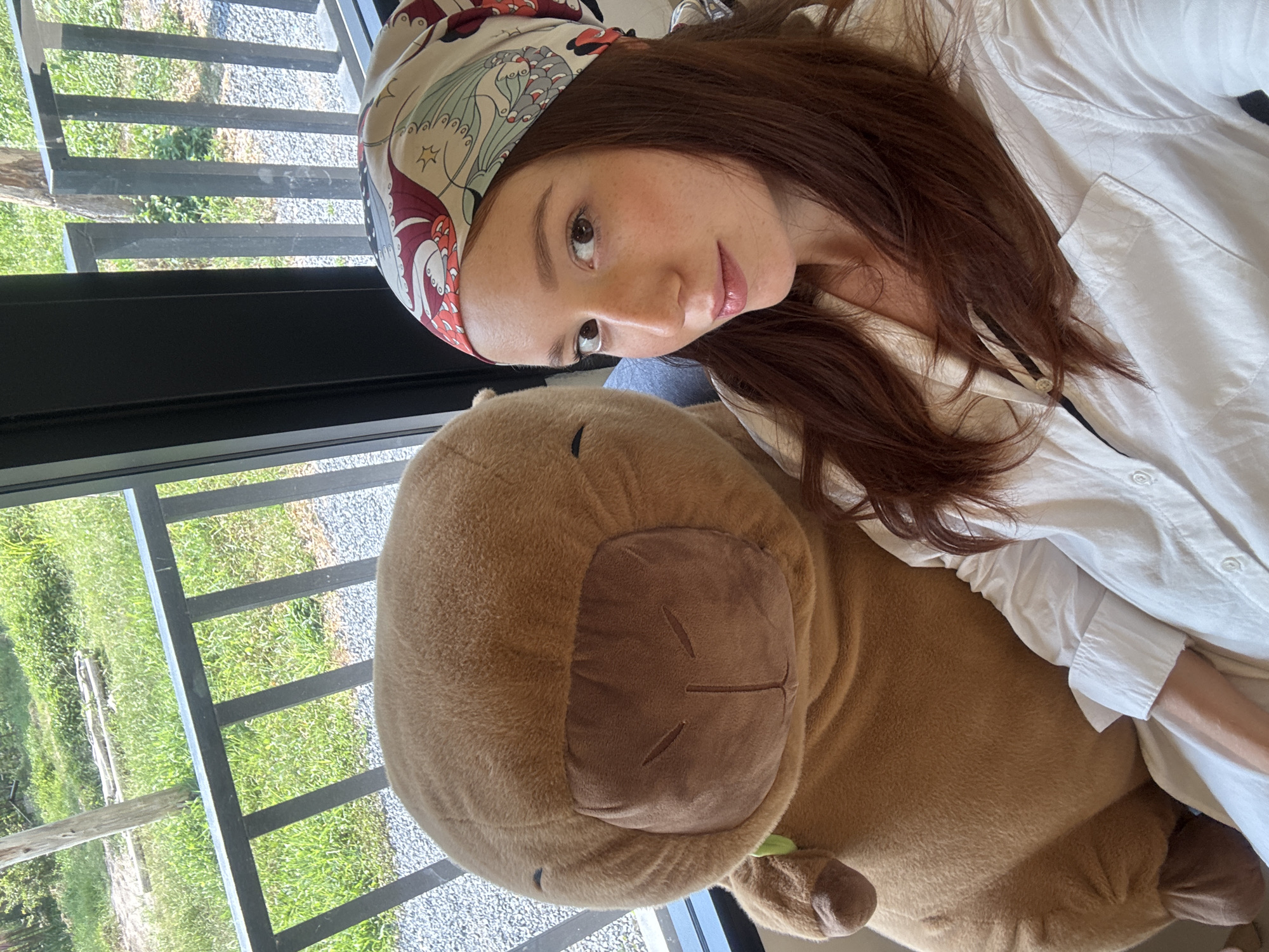 The image displays a woman and a large, brown plush toy, framed as if viewed from above or with the subjects reclined. The woman, with long, reddish-brown hair, has noticeable freckles and wears a white collared shirt and a distinctive patterned headscarf featuring colorful designs, possibly of birds or butterflies. She looks softly towards the viewer with calm brown eyes and a subtle smile.
Adjacent to her is a sizable, plush toy, resembling a capybara, in a warm brown color. It has a lighter brown snout area with simple stitched features for eyes, nose, and mouth, conveying a peaceful expression. A small green detail is visible near its lower part.
The setting appears to be indoors, next to a large window. Through the window, an outdoor scene is visible, characterized by vibrant green grass, patches of light-colored gravel, and dark, possibly metallic, railing structures. The outdoor environment suggests a natural or semi-rural landscape, consistent with locations like Choeng Thale, Thailand. The bright, natural light streaming through the window indicates daytime. The overall atmosphere of the image is relaxed and serene. There is no visible text in the media file.