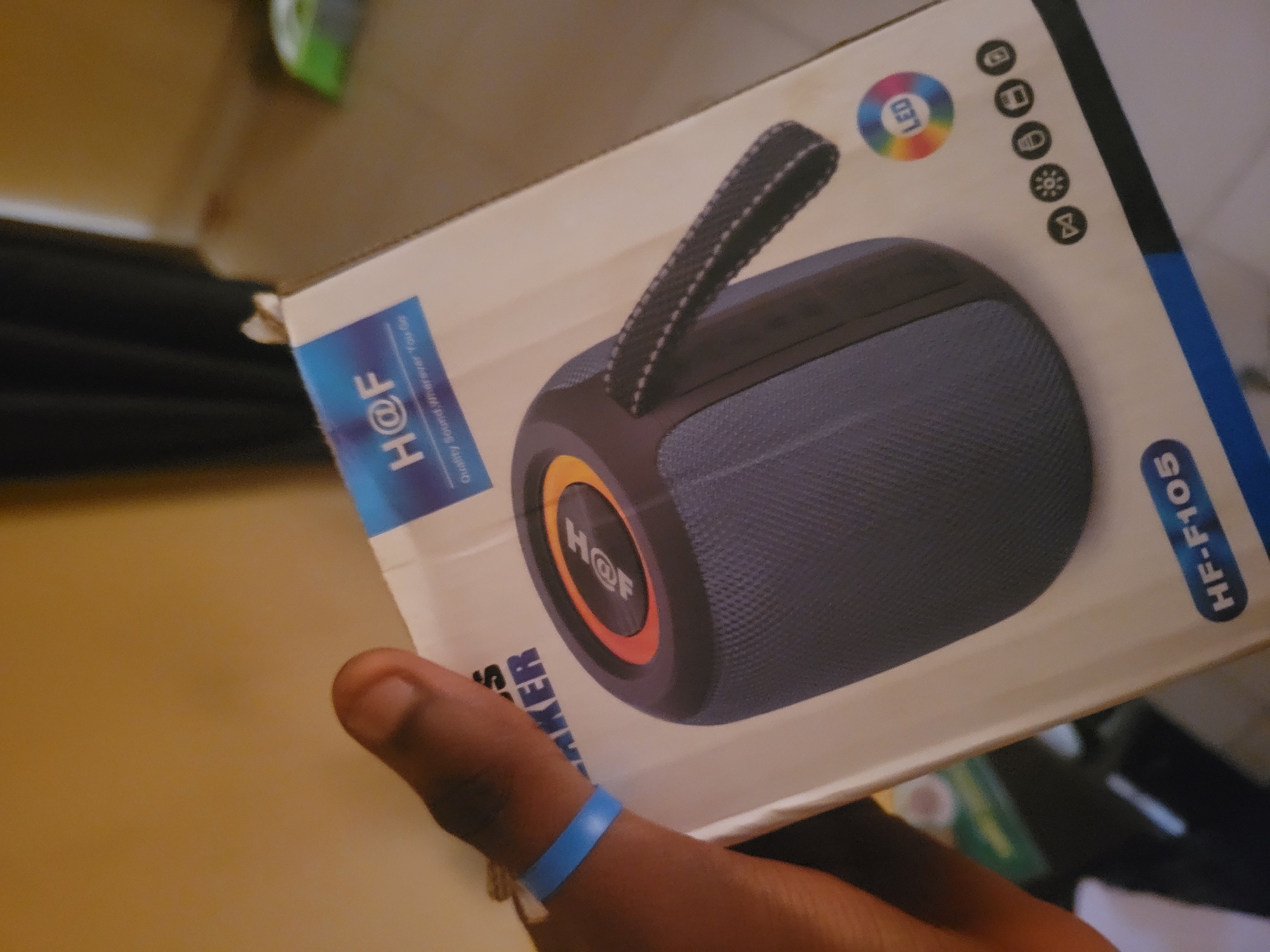 A close-up shot shows a person holding a blue wristband on their left wrist, with their thumb and forefinger partially obscuring a cardboard box. The box is white with blue and orange accents and features an image of a dark blue, cylindrical speaker with a black handle. The speaker has a circular logo with "H@F" inside, outlined in orange and yellow. Text on the box reads "H@F Quality Sound, Wherever You Go," and "HF-F105." The background is a blurry, warm-toned wall. Icons suggesting features like LED lights, battery life, and connectivity are visible on the bottom right of the box.