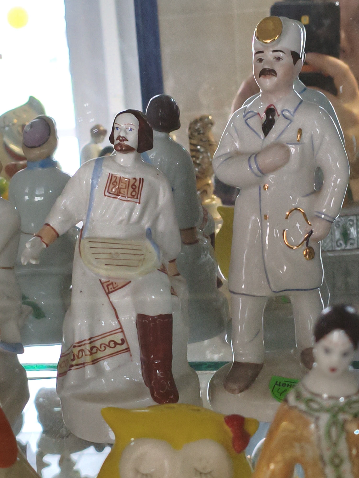 This image shows a collection of porcelain figurines displayed behind a glass surface, likely in a museum or shop in Orenburg, Russia, given the presence of a green price tag with Cyrillic text "ЦЕНА" (PRICE). The primary subjects are several human figurines, including a male doctor wearing a white coat and stethoscope, and another male figure in traditional clothing with a red trim and a satchel. In the background, other figurines are visible, including one resembling a tiger. The lighting suggests an indoor setting, possibly with natural light from a window. The overall scene is a still life arrangement of decorative items.