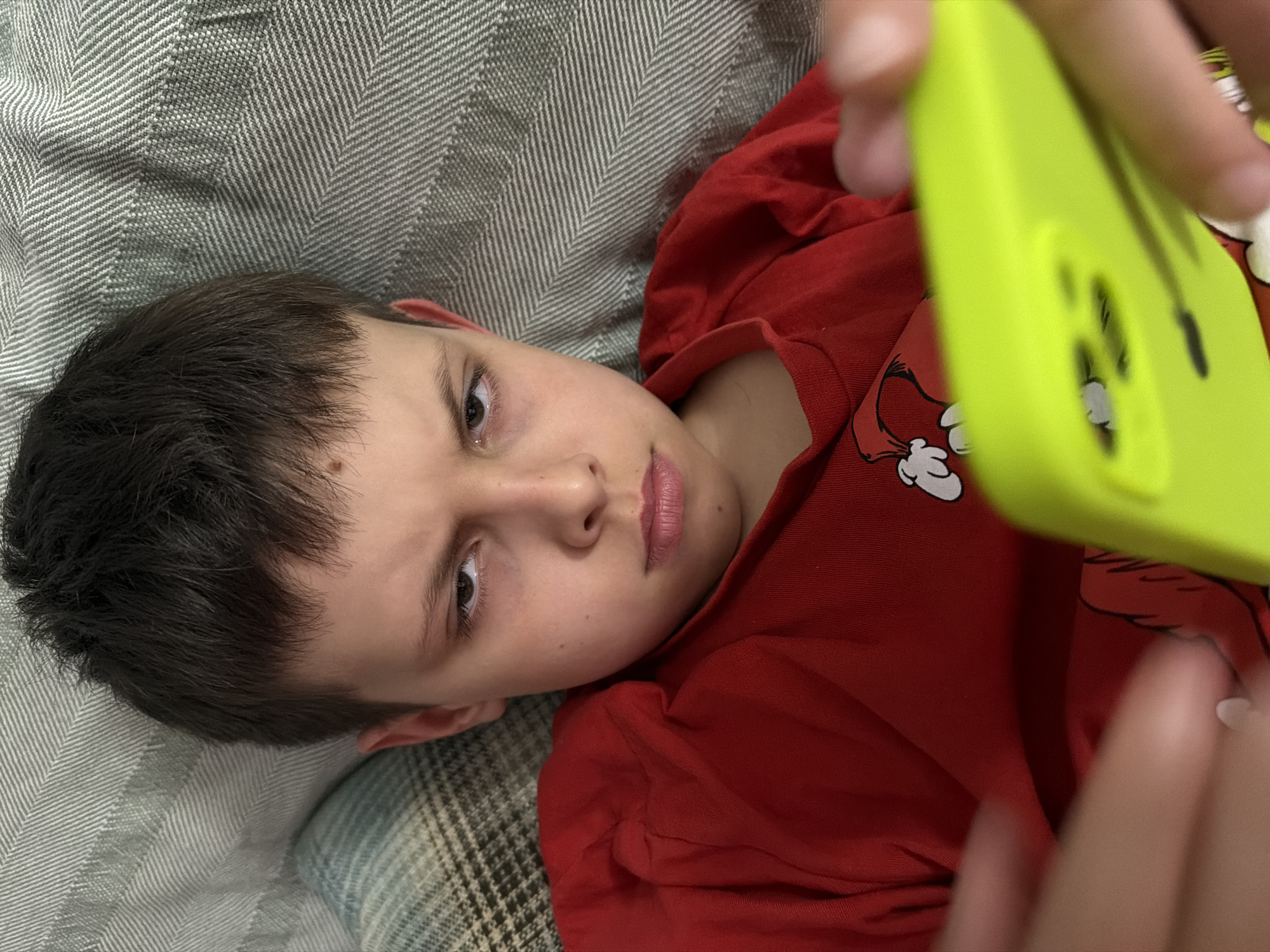 A young person lies on a herringbone-patterned gray cushion, holding a bright lime-green cell phone with their fingers visible in the lower right corner of the frame. The person is wearing a red t-shirt with a white cartoon character design. The background is blurred, but the cushion seems to be part of a larger piece of furniture, possibly a couch or bed. The location of the scene in Votkinsk, Russia cannot be ascertained from the image.