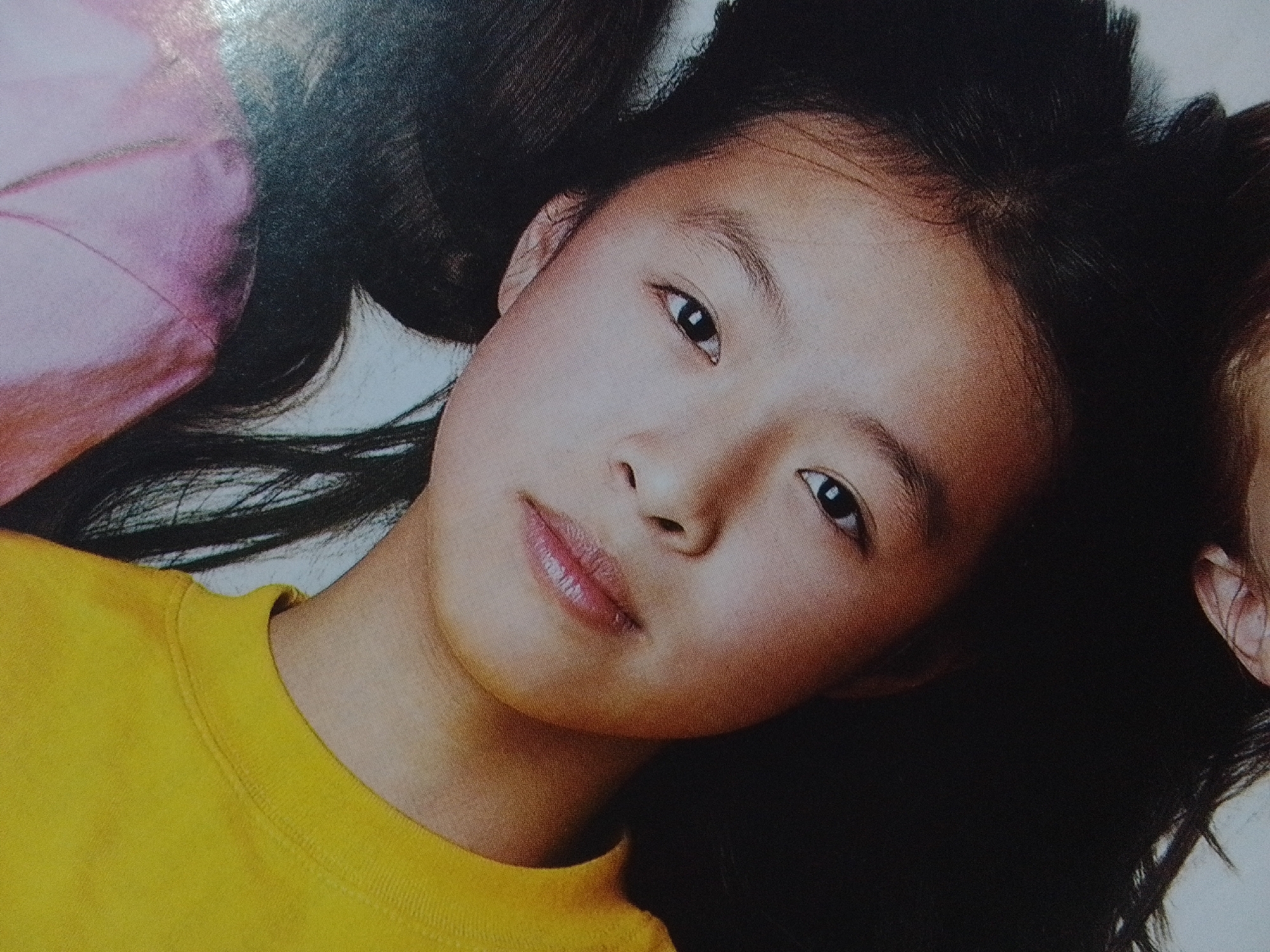 An overhead close-up shot shows a young girl lying down, her head turned to the right. Her dark hair is spread out around her head. She is wearing a yellow shirt. Her left eye is visible and looking at the camera. Her lips are slightly parted, revealing a hint of her teeth. The background is dark and out of focus. The image is a photograph of a person, not a depiction of a specific location in La Tagua, Venezuela.