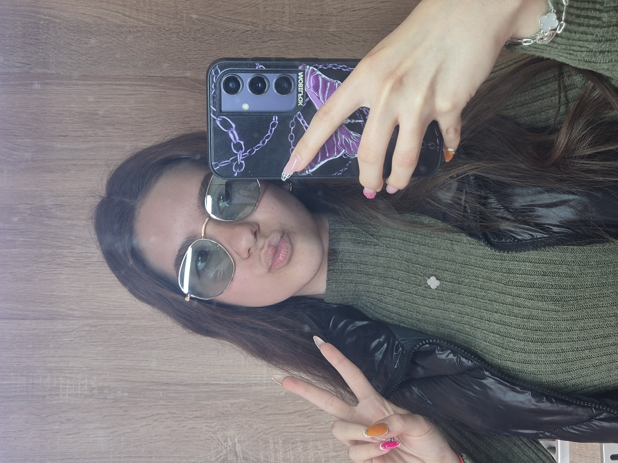 A young woman is captured in a selfie reflection, likely in Pavlikeni, Bulgaria. She has long, dark brown hair and is wearing round sunglasses with gold frames, a green ribbed knit sweater, and a shiny black puffer jacket. She holds a black smartphone with a distinct purple and black chain-patterned case, featuring a butterfly/moth design and the mirrored text "WOBITLOX," in her right hand. Her nails are long and manicured with varied designs, including pink, orange, and clear/white tips. A silver chain bracelet with a white charm is visible on her right wrist, and a similar charm hangs from a necklace over her sweater. She is making a "duck face" with puckered lips and forms a peace sign with her left hand. The background is a light-colored wooden surface, suggesting an indoor setting. The image is rotated 90 degrees clockwise, presenting a casual and playful mood.