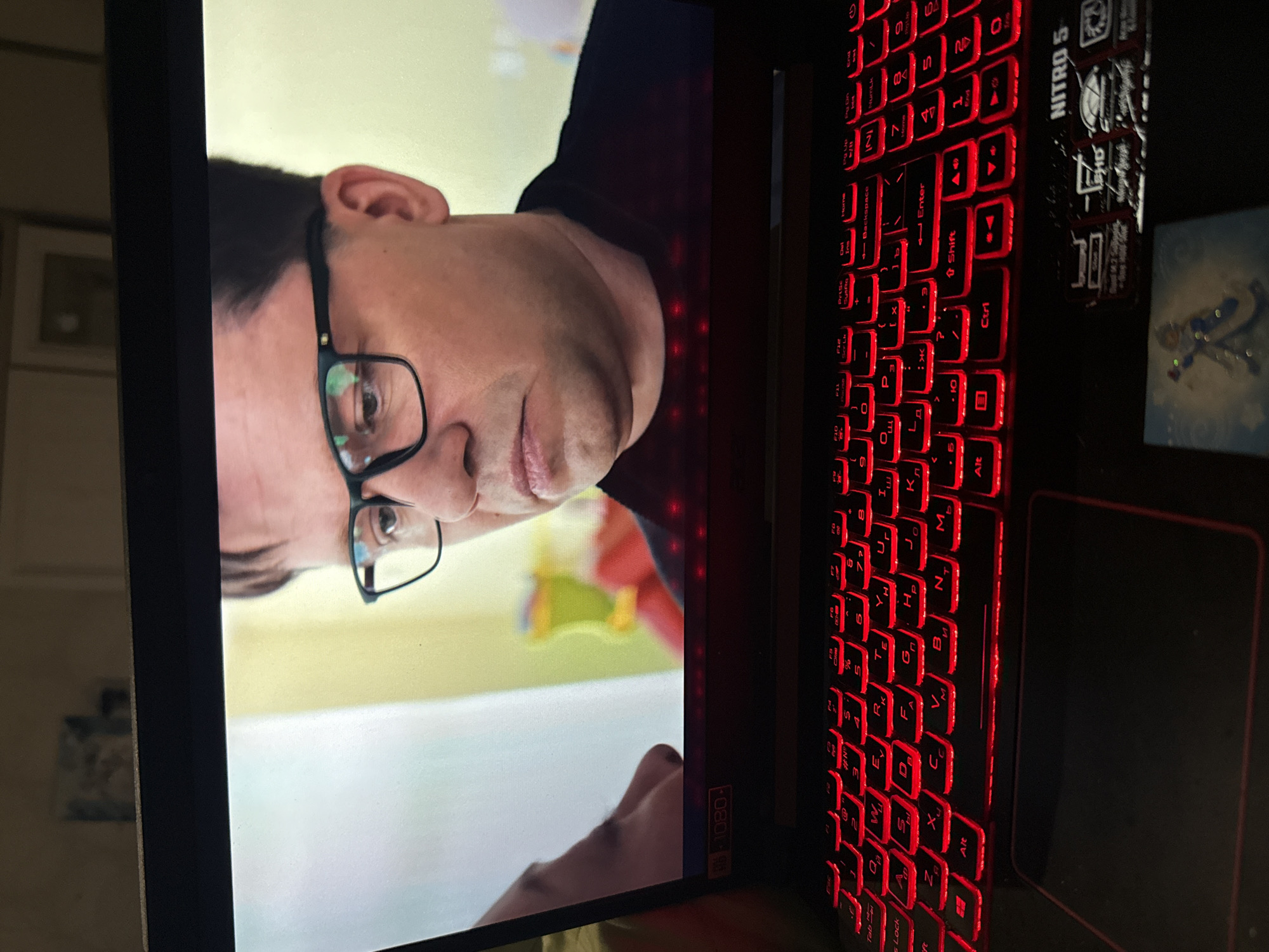 The image shows a close-up of a laptop screen displaying a man wearing glasses. The laptop has a red backlit keyboard. A sticker on the laptop indicates it is a "Nitro 5." A small, blurry item with an anime-style character is placed in front of the laptop. The background is out of focus and appears to be an indoor setting with a wall and some objects on it. The man on the screen is looking downwards, and his expression is neutral. The scene appears to be captured from a slightly high angle, looking down at the laptop.