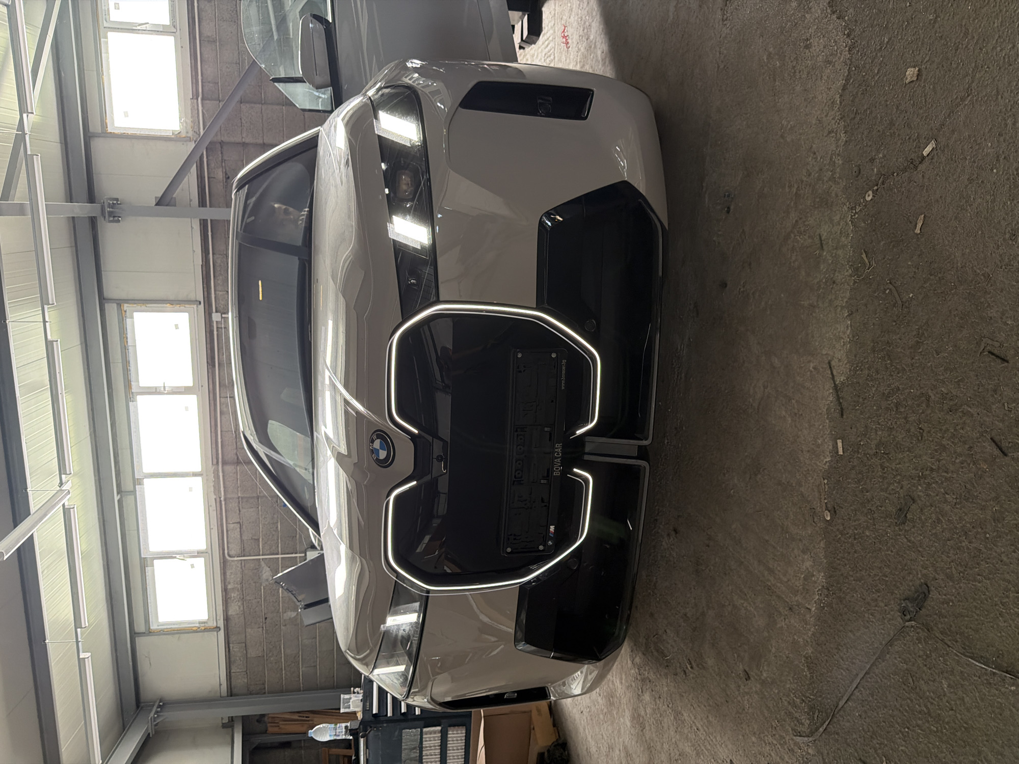 A white BMW iX is parked indoors, facing the camera. The car is in what appears to be a workshop or garage. The front of the vehicle is prominently displayed, showing its distinctive kidney grille with illuminated accents, LED headlights, and a license plate that reads "BQVA CAR". The car's doors are open. In the driver's seat, a person is visible through the windshield, looking towards the camera. The background consists of a brick wall, industrial lighting fixtures on the ceiling, and some equipment on the left side, including a toolbox and a bottle of water. The floor is concrete and appears somewhat worn. The environment suggests a commercial or service establishment.