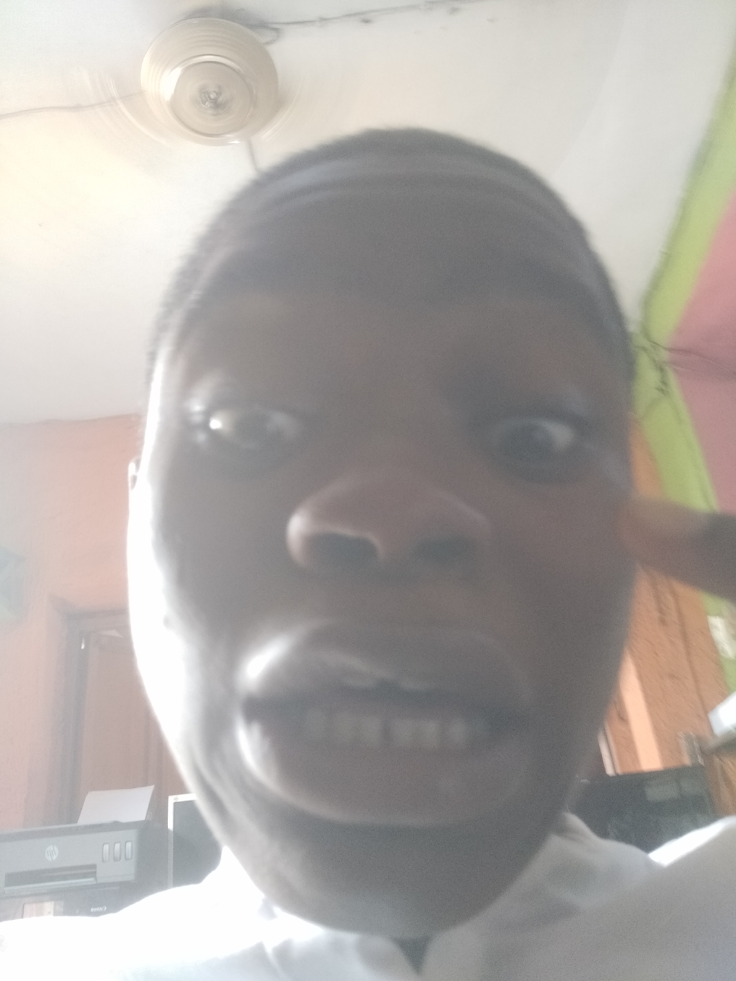 A person's face fills most of the frame, taken with a very close-up, downward angle. The person has dark skin, short dark hair, and is looking at the camera with a slightly surprised expression. The person's mouth is open, revealing their teeth.
Behind the person, the ceiling is visible, with a ceiling fan fixture. To the lower left, an HP printer sits on a desk. In the background to the right, a wall painted with green and pink is partially visible.
Based on the prompt, the location is Shangisha, Nigeria. However, there are no visible elements in the image that definitively confirm this.