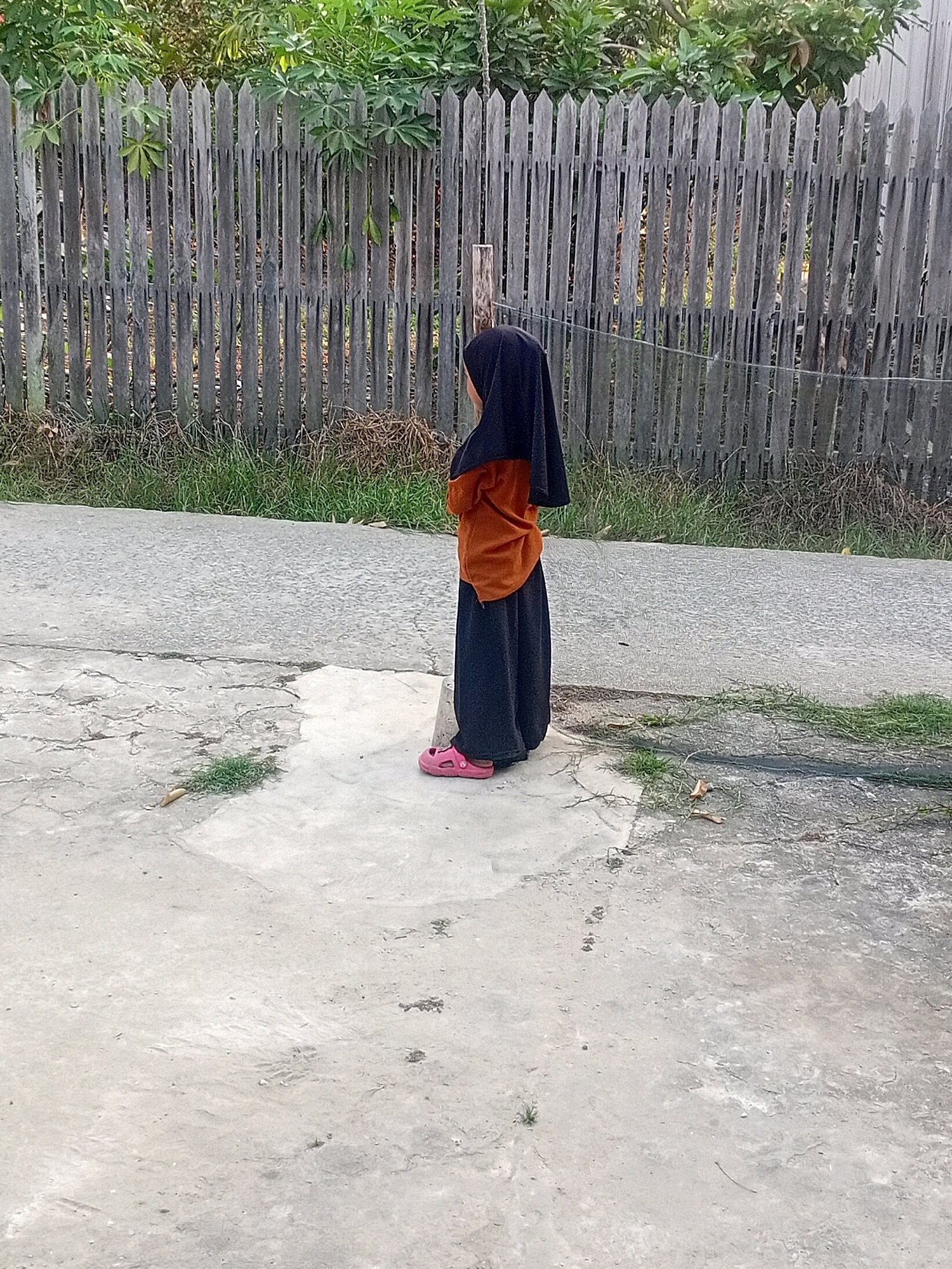 A young child, wearing a dark headscarf and an orange top with a long black skirt, stands facing away from the camera. The child is wearing pink sandals. They are positioned on a concrete surface, with a paved road or pathway to their left and a weathered wooden fence to their right. Behind the fence, lush green foliage is visible. The concrete area shows signs of wear and tear, with cracks and patches of grass growing through. The environment appears to be outdoors, likely in a residential area in Anggana, Indonesia, given the presence of the fence and vegetation. The lighting suggests it is daytime.