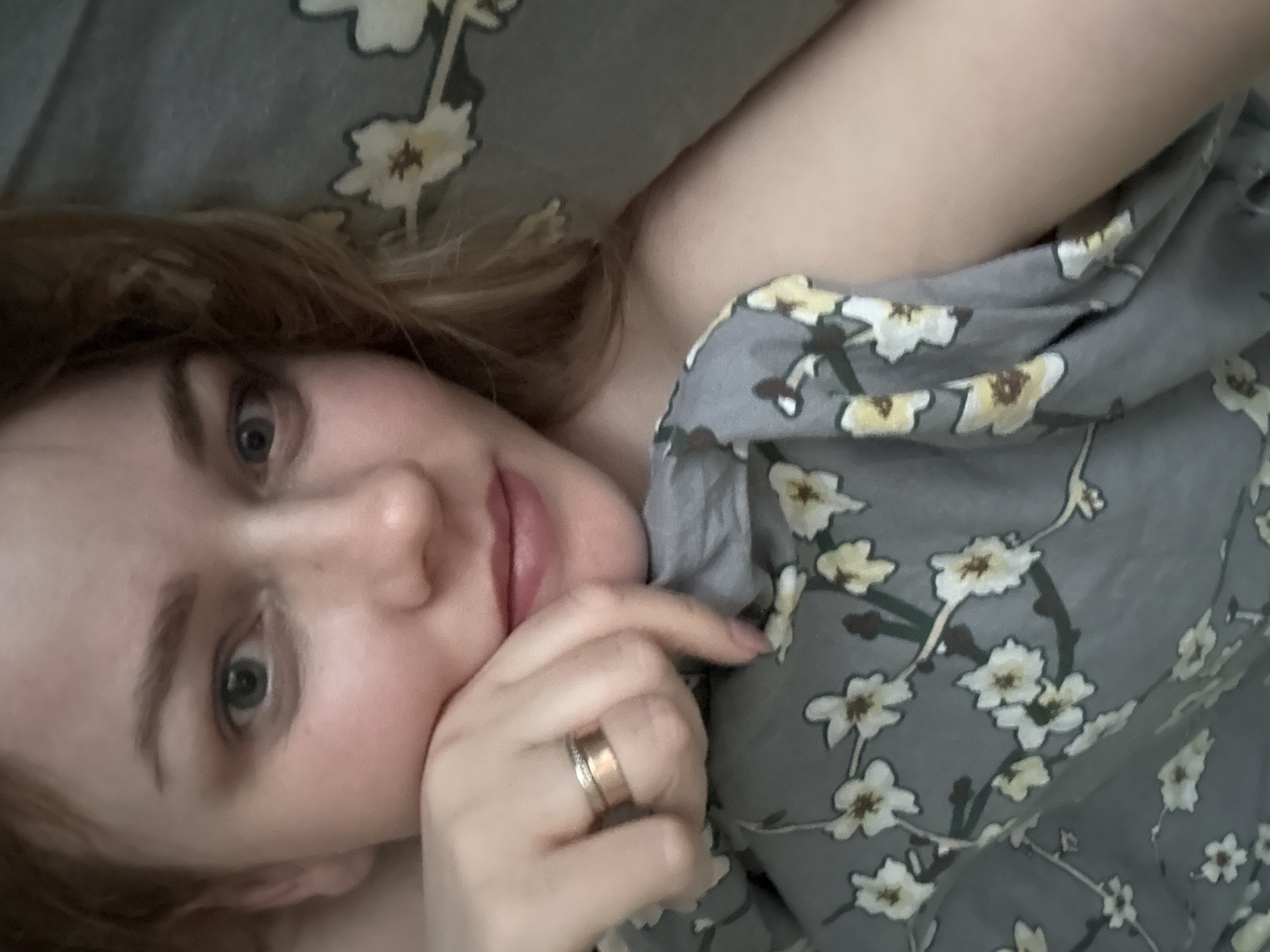 A woman is pictured close-up, lying down on a patterned surface. She rests her face on her hand, displaying a ring on her finger. The patterned surface appears to be a bed sheet or a piece of clothing with a grey background and a floral design. The lighting is soft and diffused. There is no information available to confirm the location is Chaikovsky, Russia.
