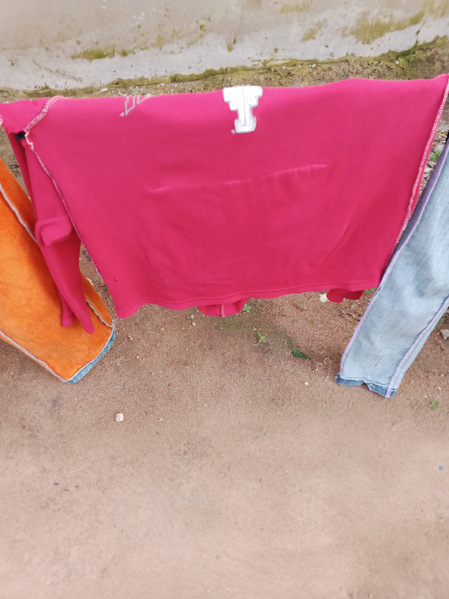 In Jauro Yinu, Nigeria, laundry hangs on a clothesline in front of a concrete wall. A bright pink garment, possibly a sweater, is draped prominently, featuring a white emblem on the upper left side. To its left, an orange towel or blanket with blue trim is partially visible. To the right of the pink garment, a pair of light blue jeans with pink stitching is also hanging. The ground below is covered in sand and scattered small stones. The overall scene depicts an outdoor drying area.