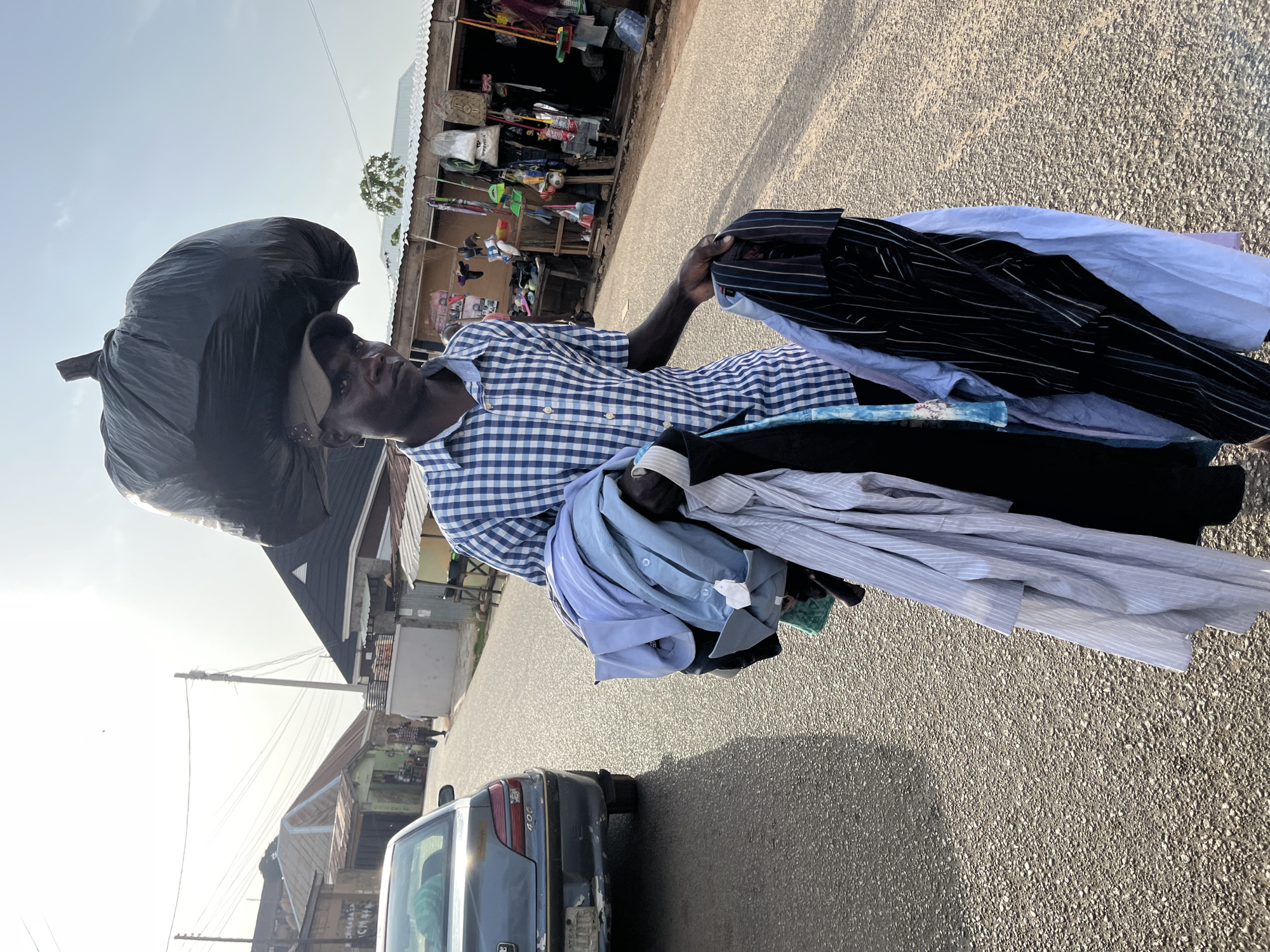 A man stands on a paved street in Jos, Nigeria, balancing a large black bag on his head and holding a stack of folded shirts in his hands. The man wears a blue and white checkered shirt and a cap. The shirts he's holding include various colors and patterns, such as a black pinstripe shirt, a light blue shirt, and a white striped shirt. Behind him, there is a storefront with various items displayed. The back of a silver car is visible in the lower left corner. Overhead electrical lines are visible against a pale sky. The scene appears to be in a residential or commercial area with buildings of varying heights.