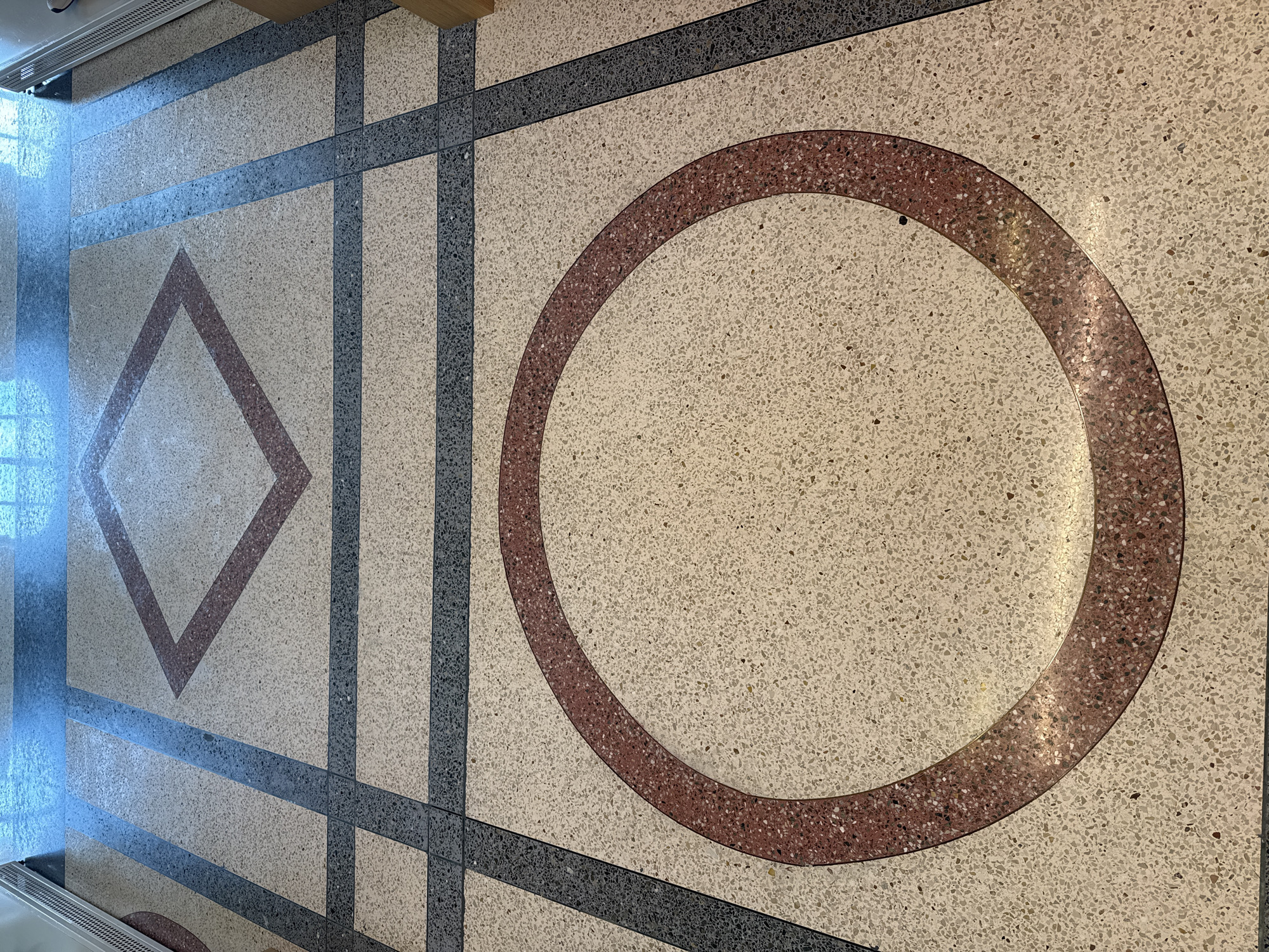 The image shows a close-up view of a terrazzo floor with geometric patterns. The floor is a light beige color with small flecks of various colors, creating a speckled texture. There are inlaid bands of dark gray and thin, reddish-brown lines forming geometric shapes. In the foreground, a large, incomplete circle of reddish-brown material is visible. Further back, a diamond shape with reddish-brown outlines is also present, intersected by dark gray bands. The flooring appears to be in a public indoor space, possibly a lobby or hallway, with light reflecting off the surface from overhead lighting. The scene is static and devoid of people. The location of this floor in Sochi, Russia, is not determinable from the provided image.