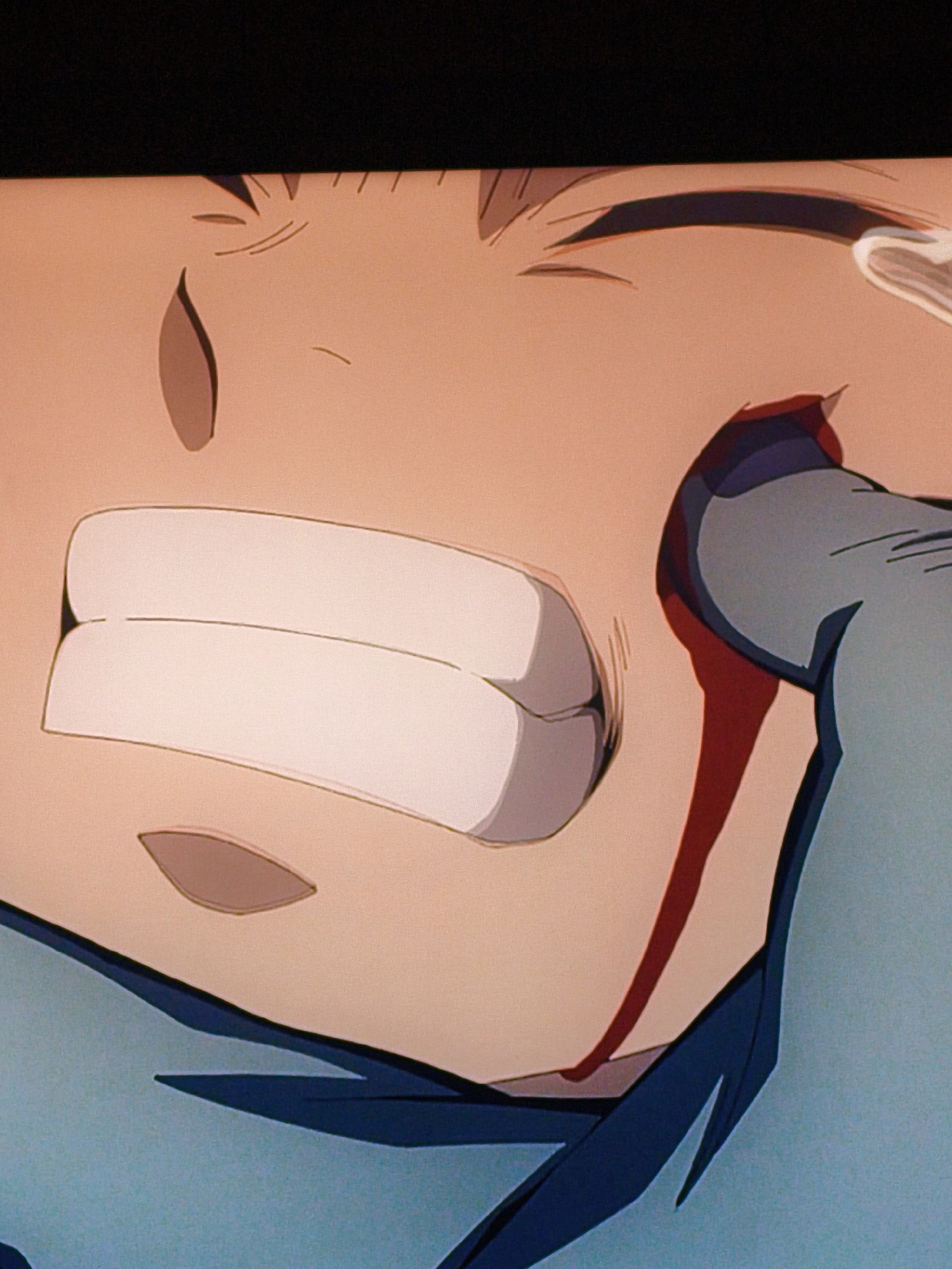 The image is a close-up, cropped shot of an anime character's face. The character appears to be a young male with light skin and dark hair, which is partially visible at the top of the frame. His mouth is open, revealing white teeth and a pinkish interior. There is a prominent streak of red, representing blood, running down from his cheekbone to his jawline. The character's eye area is visible, showing an eye with dark eyebrows and what appears to be a tear or sweat droplet on his temple. The overall color palette is warm, with flesh tones and muted blues and grays for clothing or other elements. The image does not depict any elements that would allow for identification of its location in Vladivostok, Russia, or any specific urban environment. It is solely a depiction of an animated character.