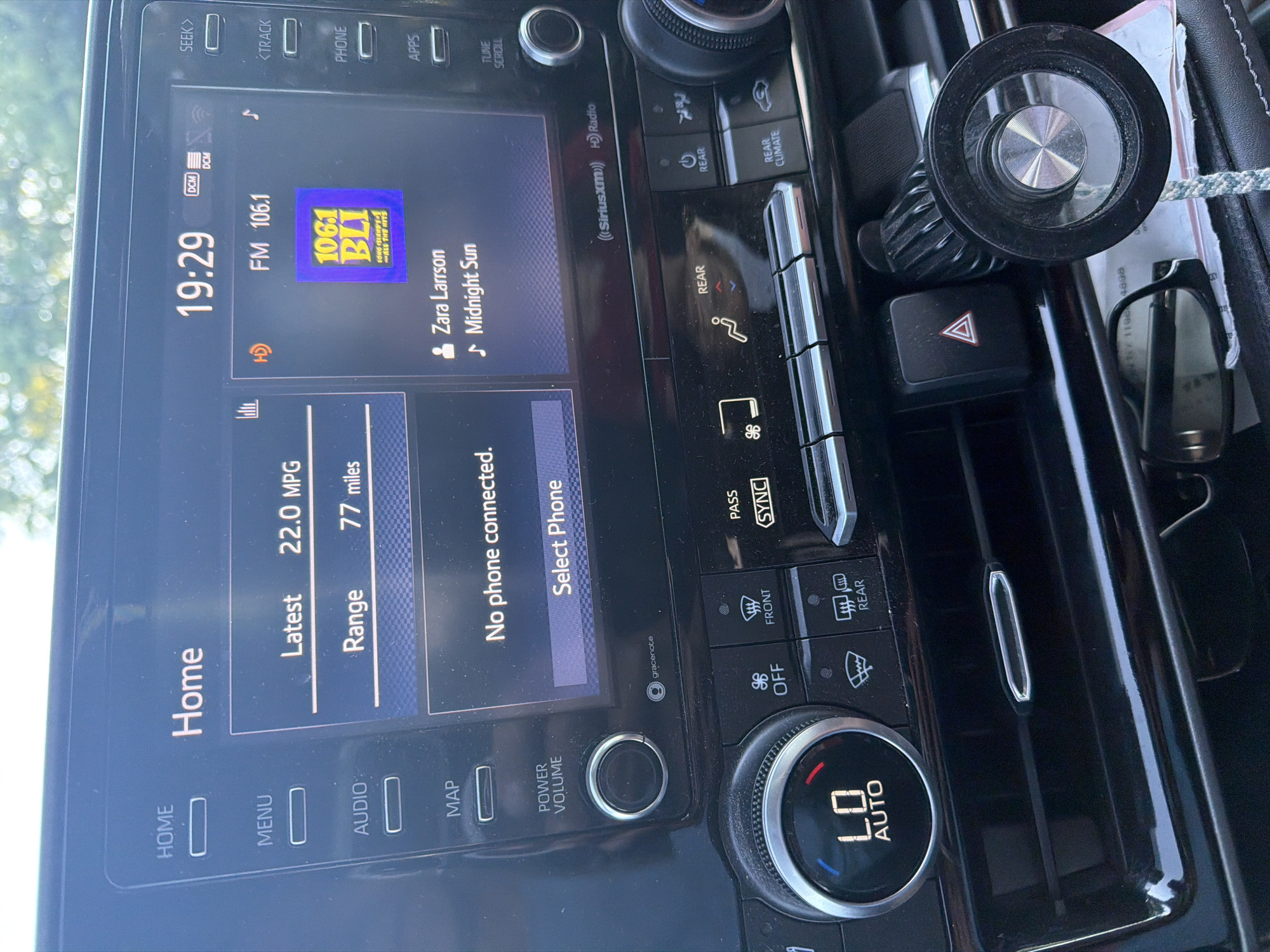 This image is a close-up, eye-level shot of a car's infotainment system and dashboard. The screen displays the time as 19:29, the radio station FM 106.1 BLI, and information about the car's fuel efficiency (22.0 MPG, 77 miles range). It also shows "No phone connected" and an option to "Select Phone." Below the screen are various buttons and knobs for controlling the car's functions, including climate control, audio, and navigation. The overall environment appears to be the interior of a modern vehicle, suggesting the car is parked or in transit. There are no people visible in the image. The specific location in Riverhead, United States of America, cannot be determined from the visual information provided.