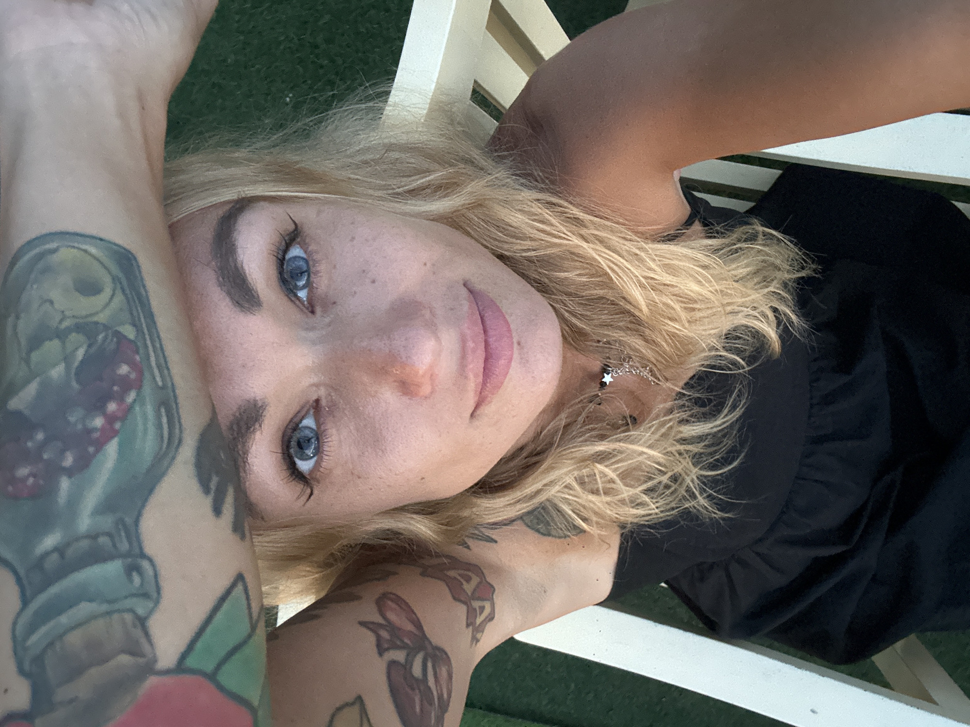 A close-up, high-angle, selfie-style photograph shows a young Caucasian woman with blonde hair and blue eyes. She is wearing a black strapless dress and a silver necklace with a star pendant. Her right arm is raised above her head, obscuring part of her forehead, and her left arm is bent at the elbow, resting on her shoulder. Both arms are covered in colorful tattoos. She is seated on a white wooden chair outdoors, with green grass visible in the background. The scene does not provide any information about the location in the city of Phu Thuy Ward, Vietnam.