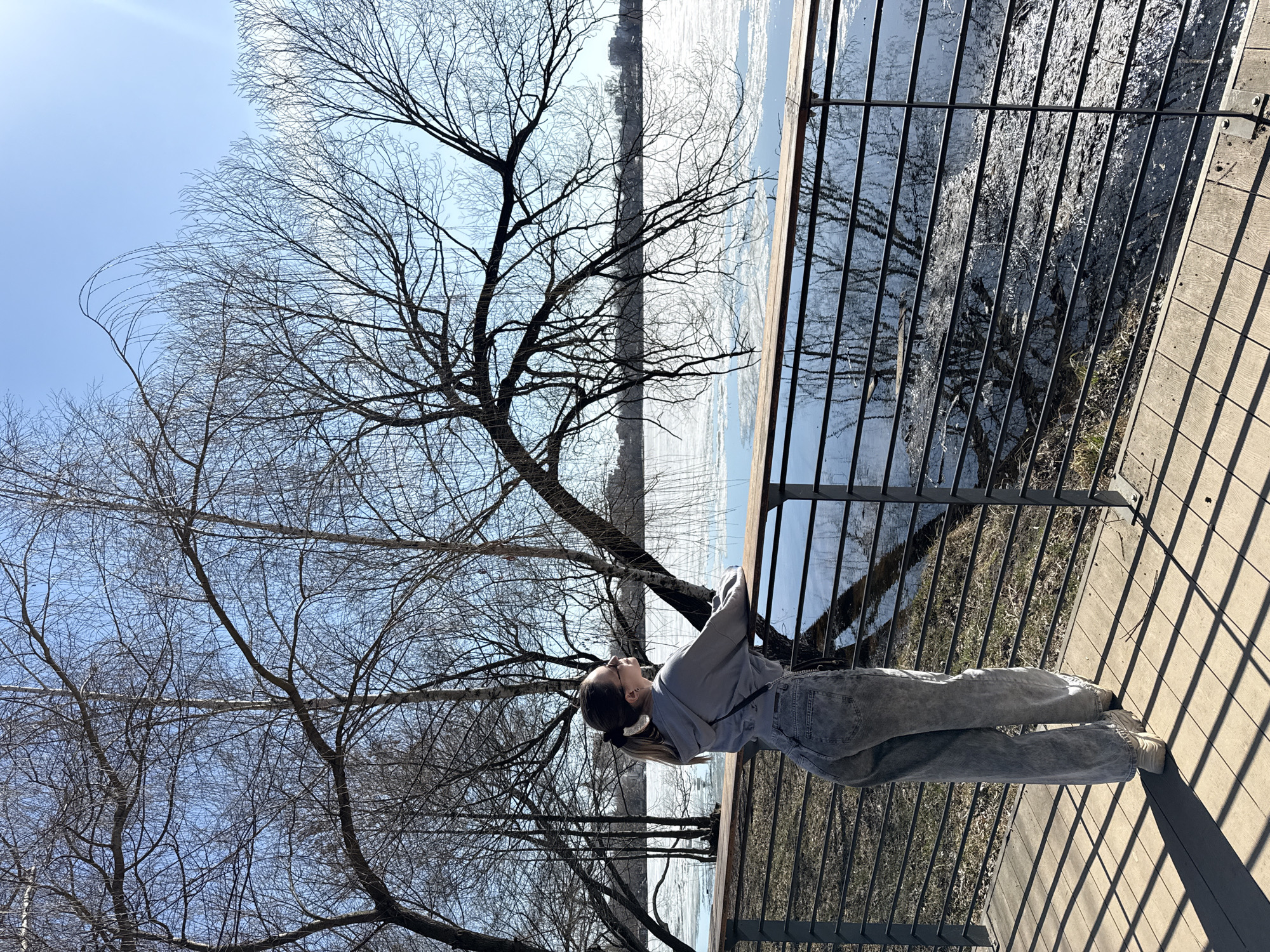 A woman is standing on a wooden platform, leaning against a black metal railing. She is wearing a gray sweatshirt, blue jeans, and light-colored shoes. She has her hair pulled back in a ponytail. Behind her, a bare tree stretches its branches against a blue sky. The scene overlooks a body of water, possibly a river or lake, with some ice visible on the surface. The location is Moscow, Russia.