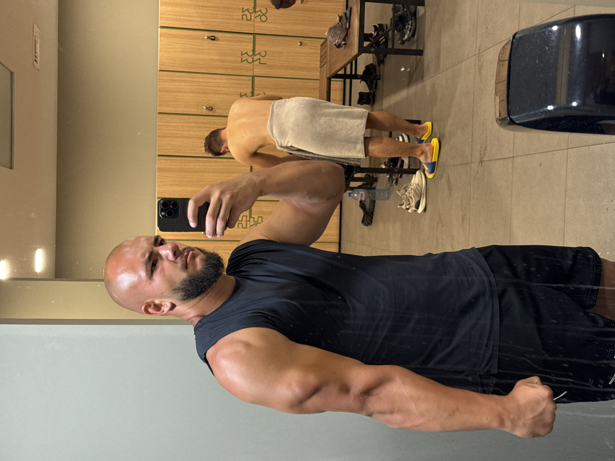 The image depicts a gym locker room, likely in Sofia, Bulgaria, based on the presence of cyrillic lettering on the lockers. The foreground features a bald, muscular man with a beard taking a selfie in a black sleeveless shirt and black shorts. His right arm is flexed, showcasing prominent muscles. In the background, another man, shirtless and wearing a grey towel, is visible. The locker room has wooden lockers, benches, and tiled flooring. Various gym-related items like shoes, a water bottle, and a hand dryer are also present. The overall scene suggests a post-workout environment.