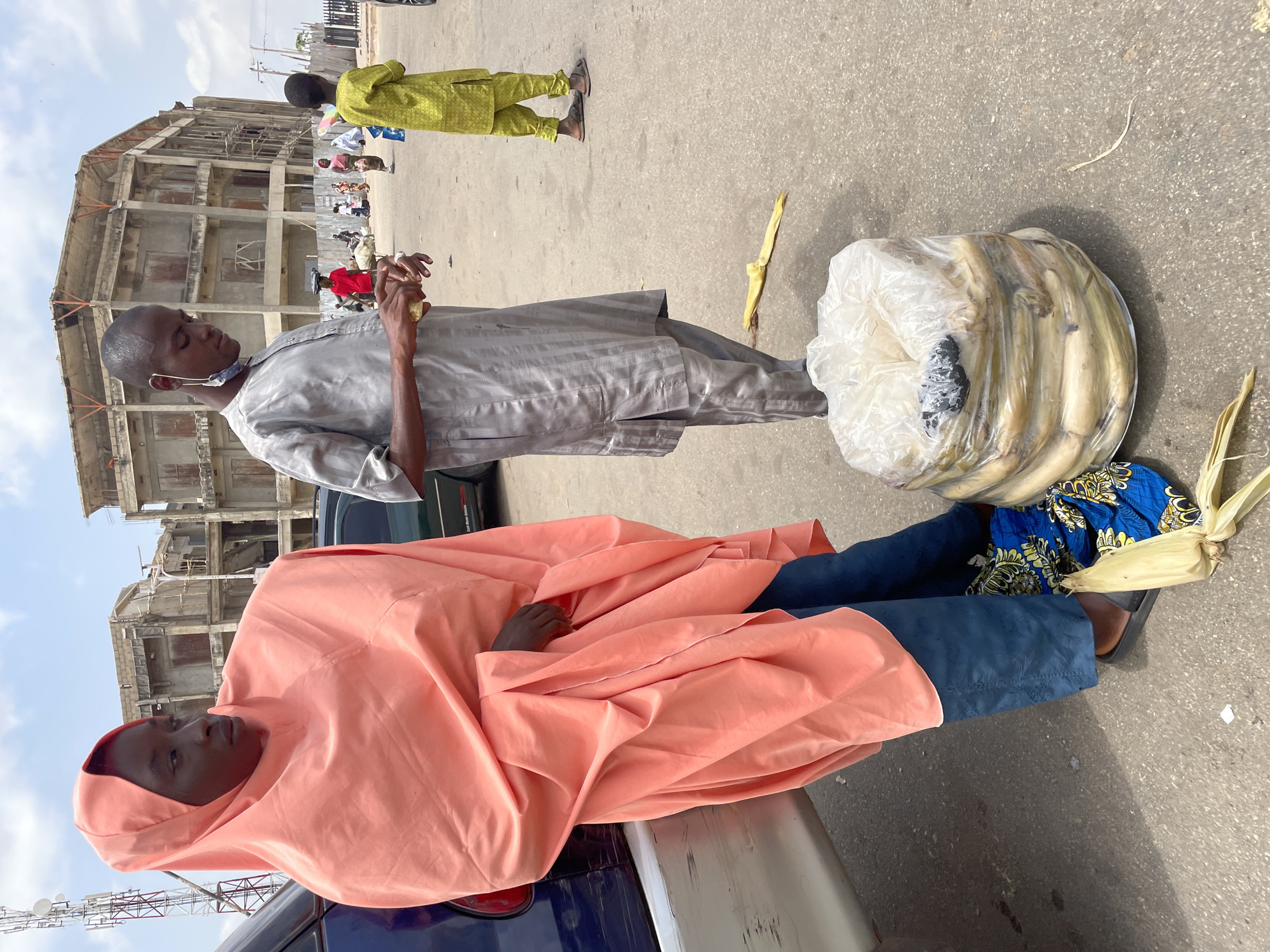 Two people are pictured outdoors near an unpaved city street. One person is reclining on a dark blue vehicle and is draped in a peach-colored robe. The other person is standing, wearing a gray tunic and pants with a face mask hanging below the nose, and holding food in the hand. There is a pile of corn in a clear plastic bag next to the standing person. The background includes a building under construction and other pedestrians. The location is in Jos, Nigeria.