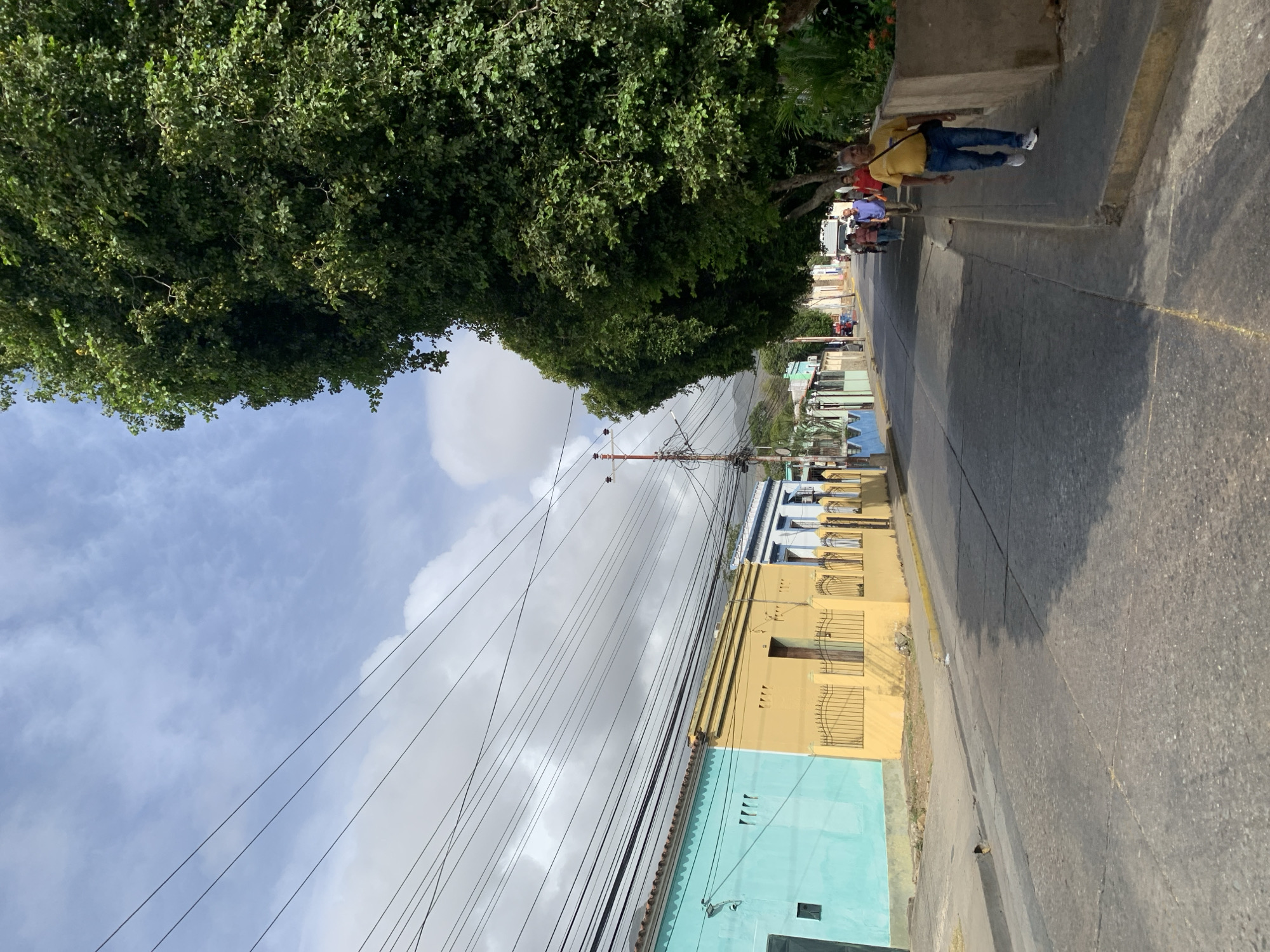 A street scene in La Asuncion, Venezuela, shows a concrete sidewalk and a paved street running through a neighborhood. Overhead power lines stretch across the sky, partially obscured by a large tree canopy. Buildings painted in light blue and yellow line the street. Several people walk along the sidewalk. One individual wears a yellow shirt and blue jeans.