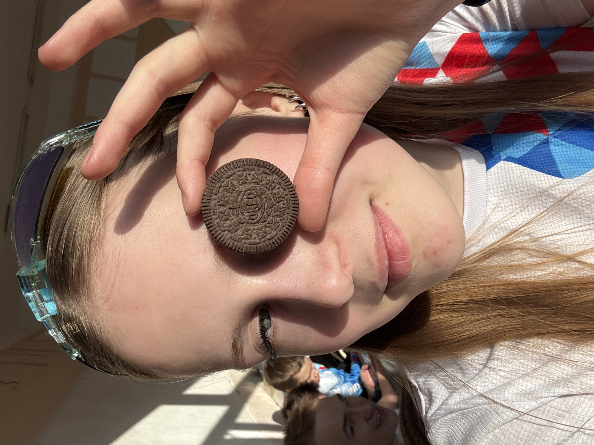 A person is holding an Oreo cookie over their eye, with their fingers framing the cookie. The person is wearing sunglasses and a white t-shirt with a red, white, and blue design. The image is taken in an outdoor setting with sunlight. The background is out of focus, but it appears to show other people. The specific location within Yuzhnyy, Russia, cannot be determined from the image.