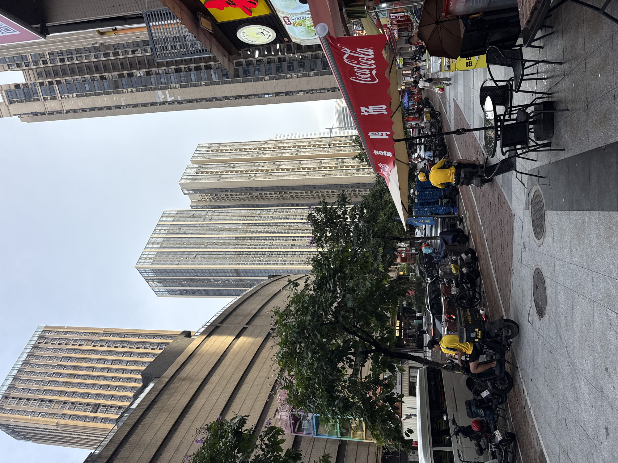 This is a street scene in Haizhu District, China. The image captures a busy street with tall, modern apartment buildings lining one side. In the foreground, a sidewalk is lined with trees and a row of parked motorcycles and scooters. A delivery person wearing a yellow shirt and helmet rides a scooter. To the right, a street-level food stall is set up under a red awning that says "Coca-Cola" in English. There are tables and chairs set up outside the stall. The street appears to be paved with grey stone tiles. The sky is overcast.
